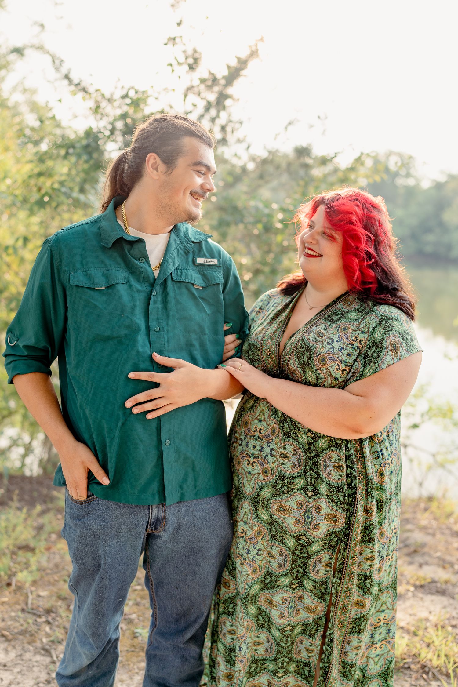 A couple shares a candid laughing moment together outdoors wearing green and floral clothing in natural lighting.