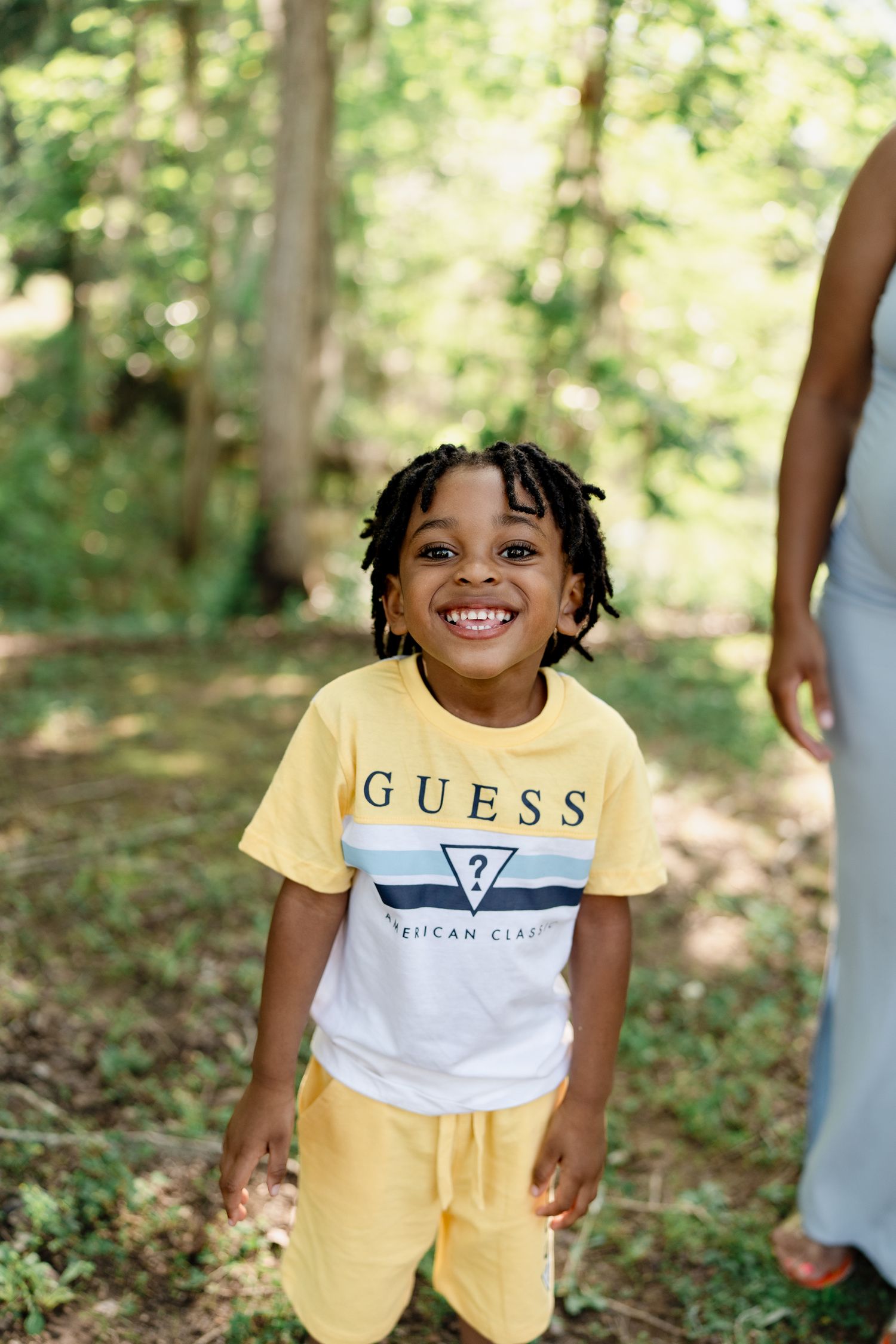 Young child wearing yellow and white Guess t-shirt smiling in wooded outdoor setting.