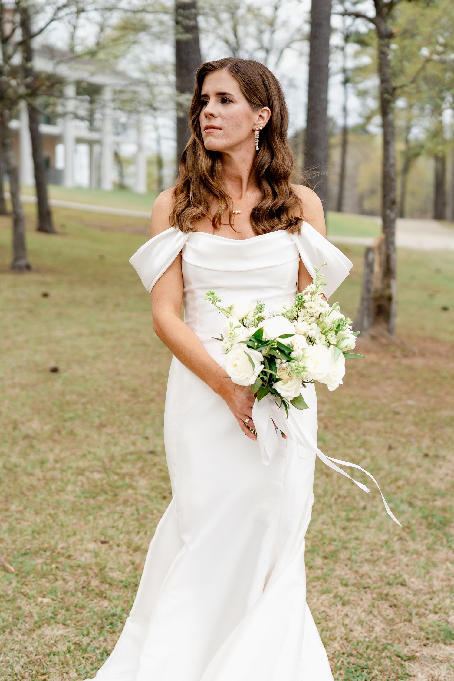 Elegant bride in off-shoulder wedding gown holds white bouquet against pine forest backdrop.