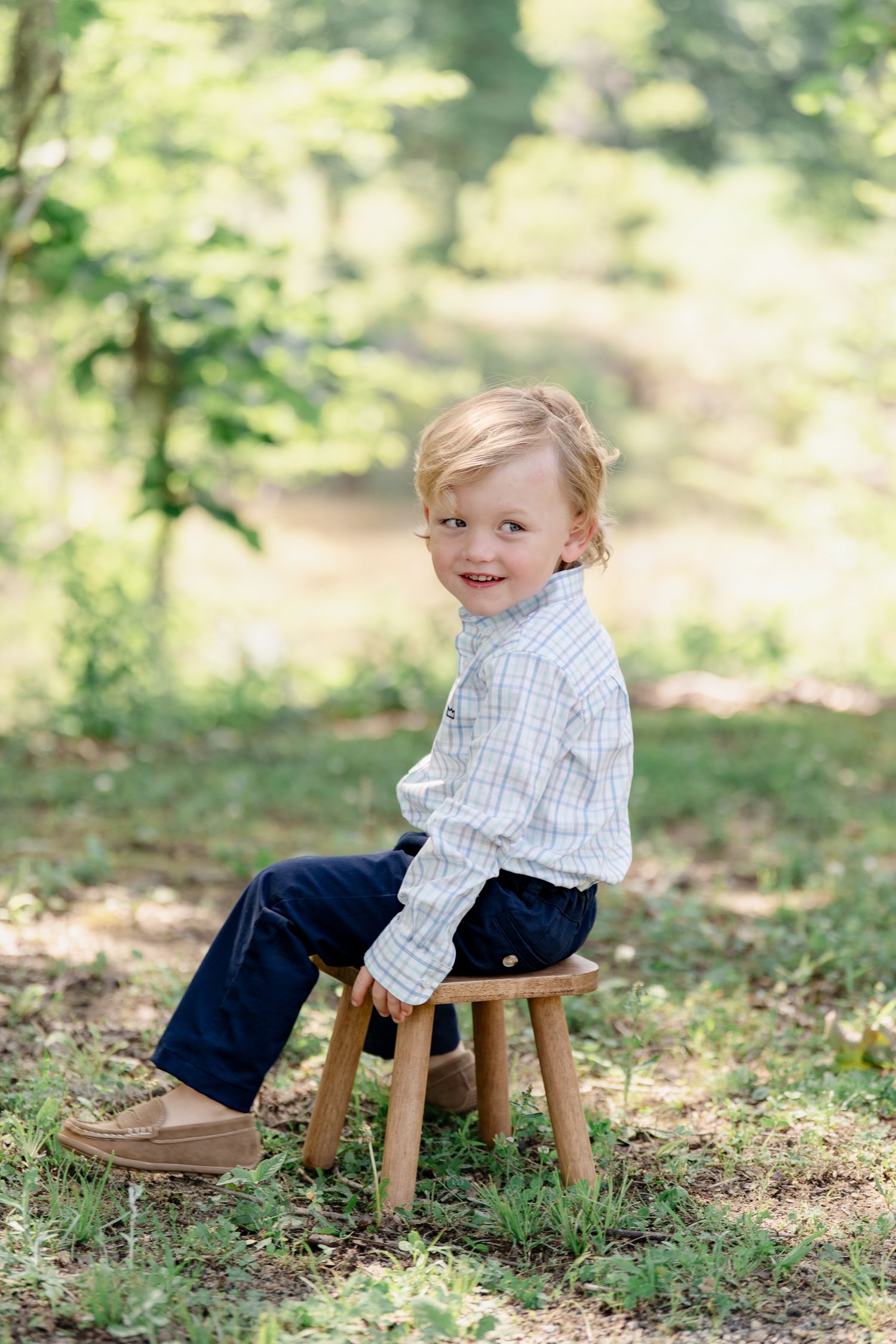 Child sitting on wooden stool for portrait session in dappled forest light.