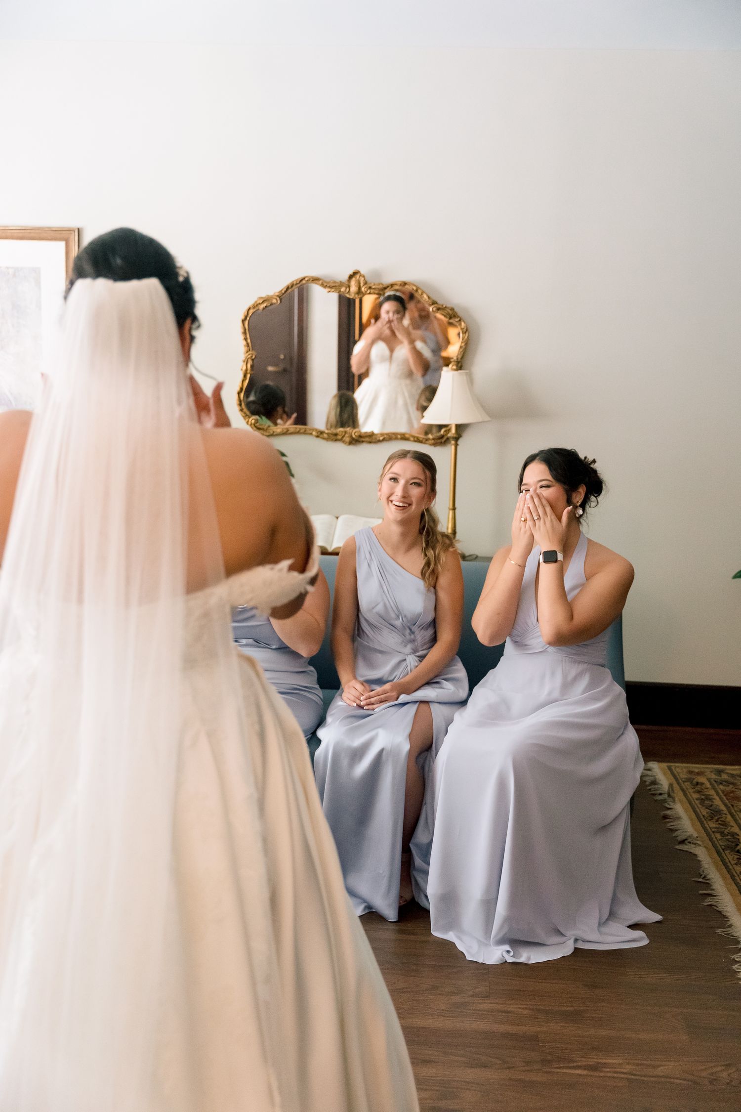 Bridesmaids in light blue dresses react emotionally to seeing the bride in her wedding gown.