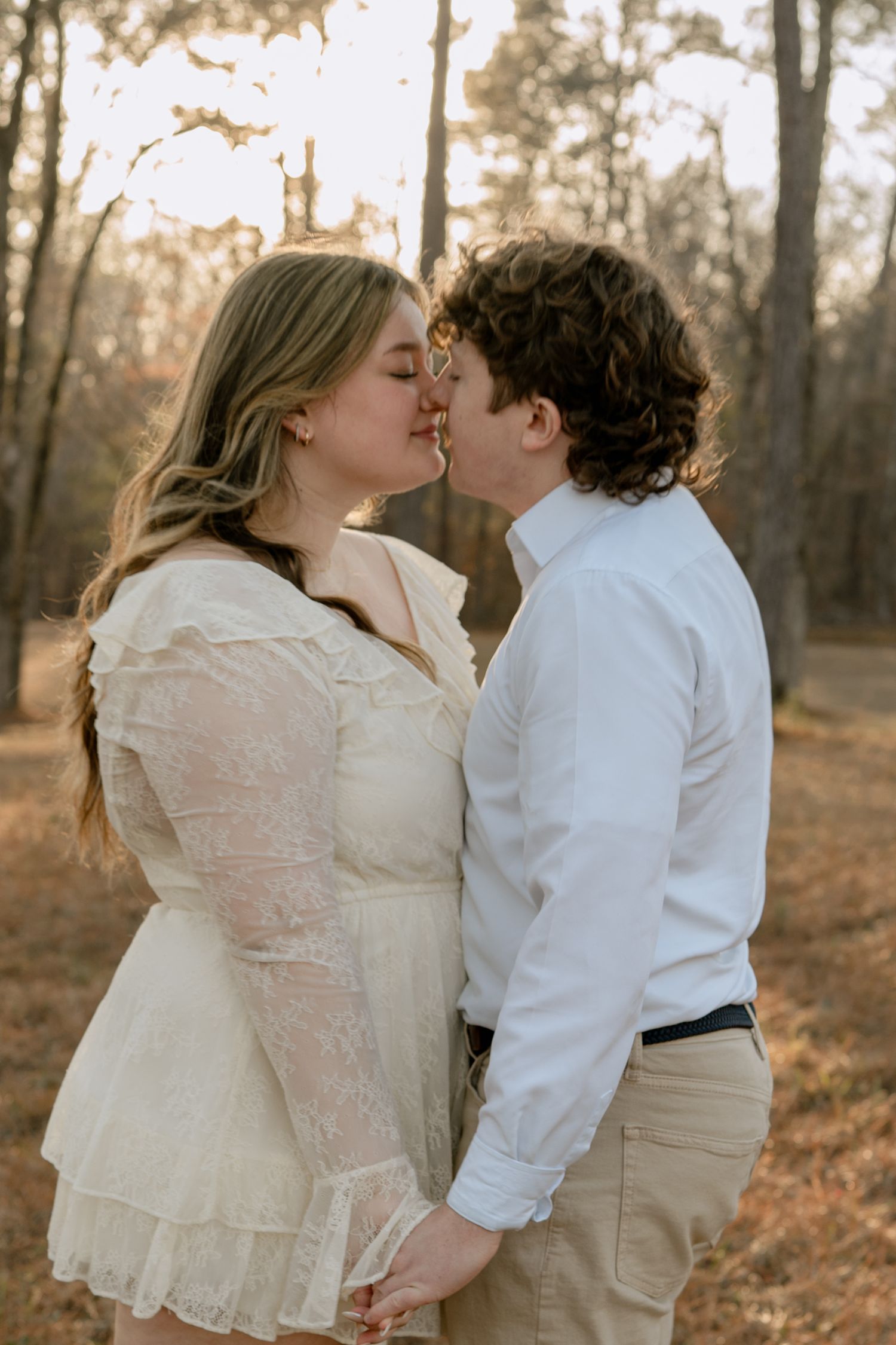 Couple sharing a kiss while holding hands in a sun-drenched pine forest during golden hour.