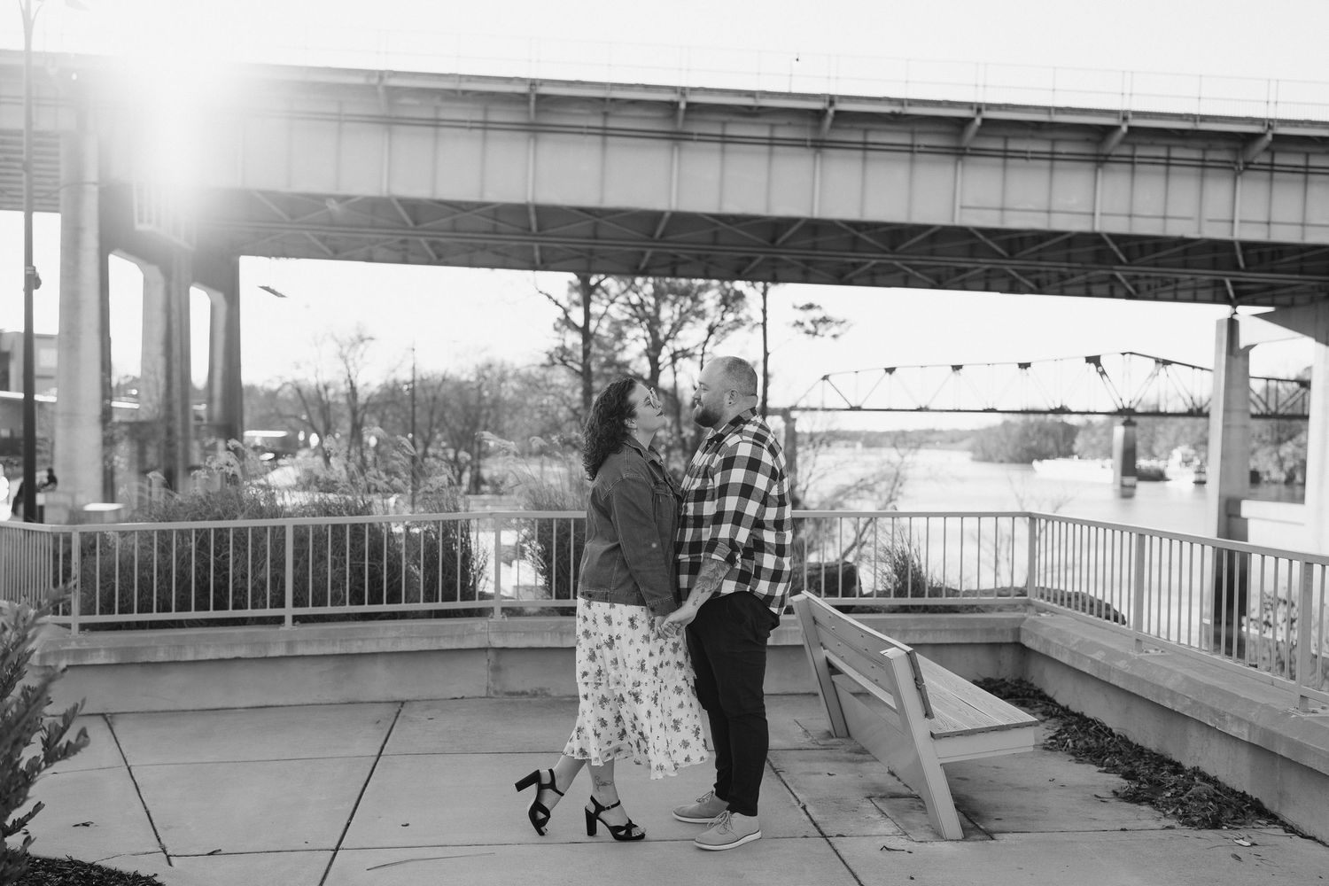 A couple stands together under a bridge structure near the water on an overcast day.