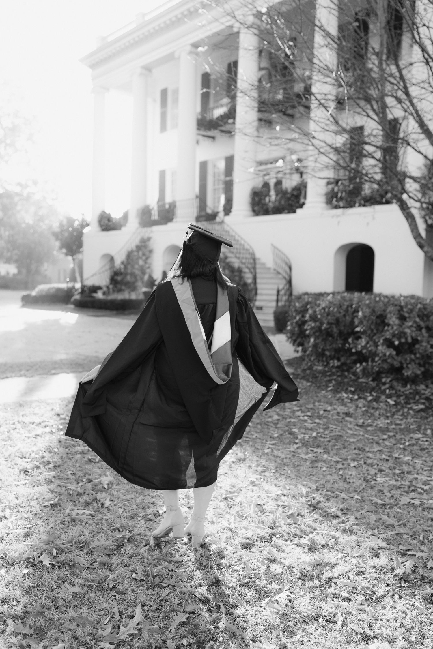 Black and white photo of graduate twirling in academic regalia on leaf-covered lawn.