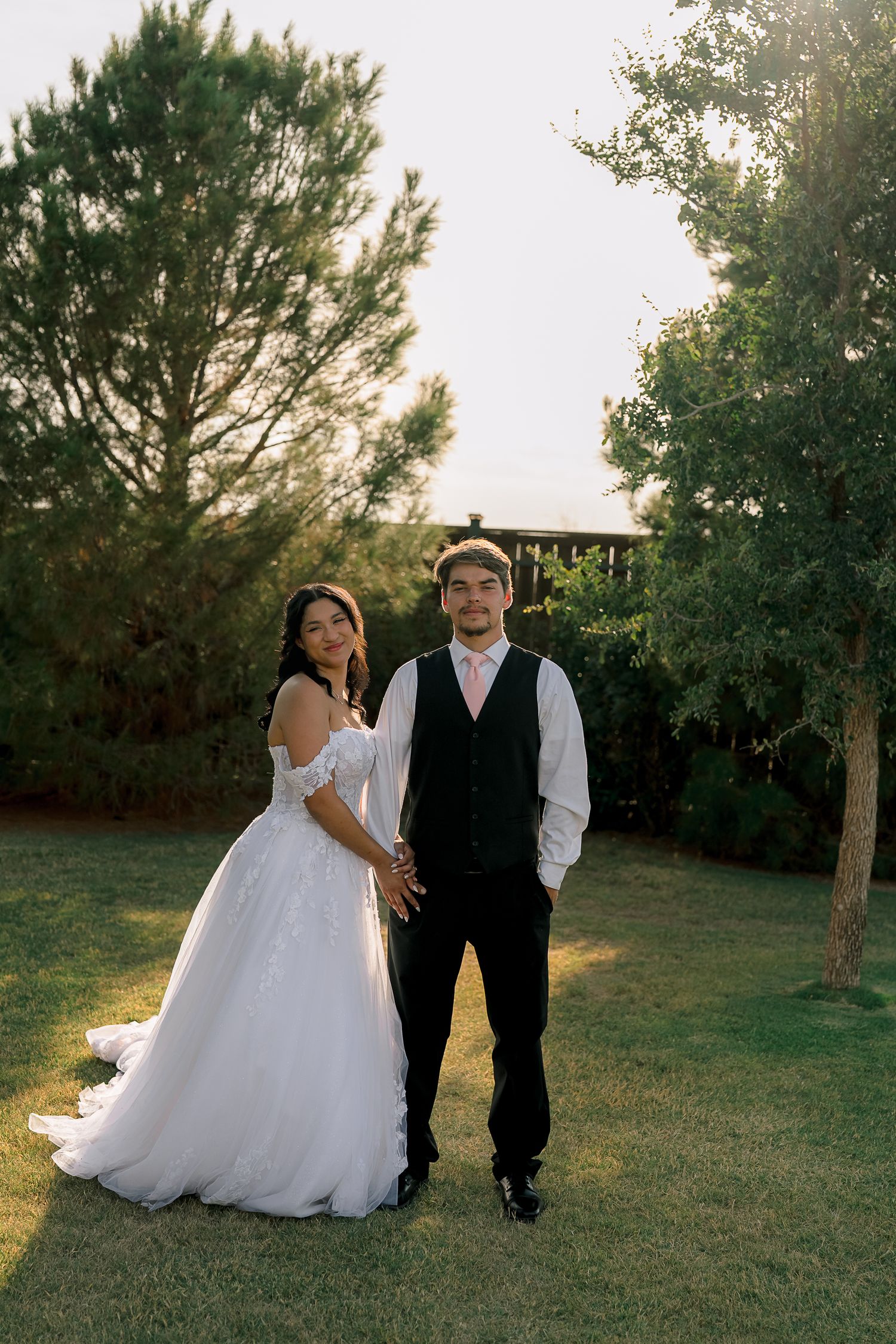 Bride in elegant white gown and groom in black vest holding hands in a lush green garden at sunset.