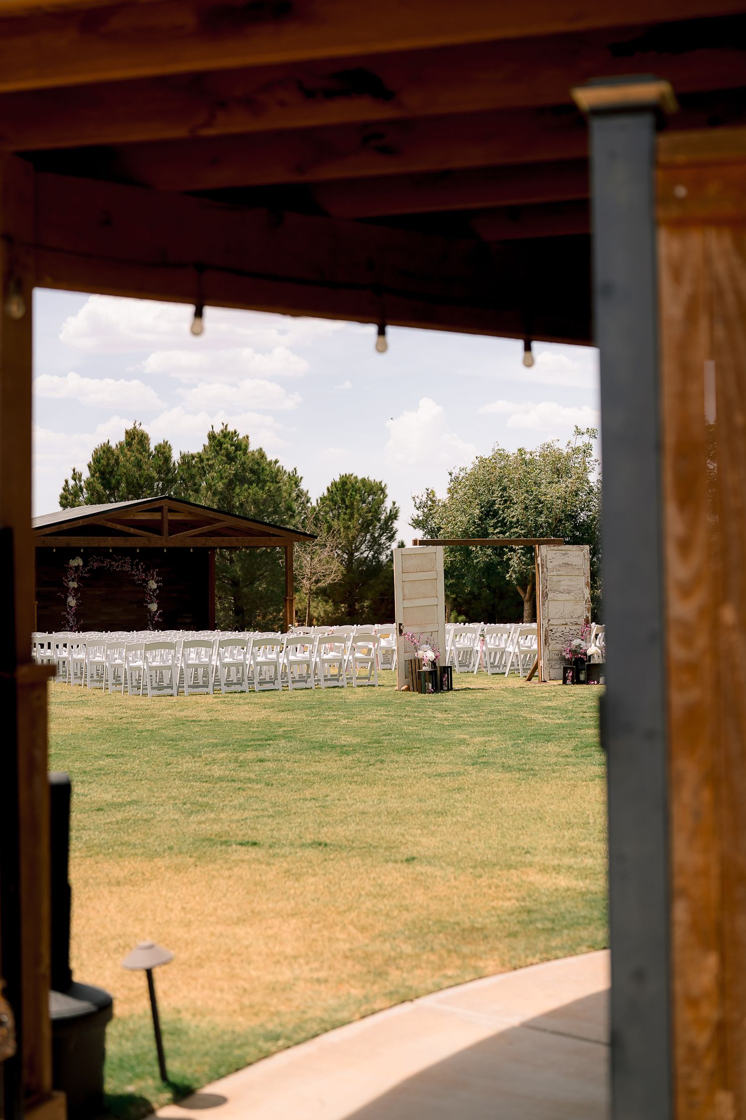 Outdoor wedding ceremony setup with white chairs on green lawn viewed through rustic wooden barn entrance.