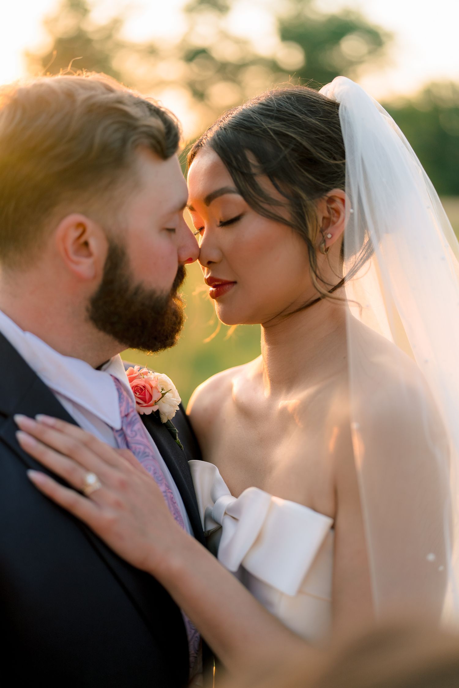A bride and groom share a romantic golden hour moment, noses touching, as sunlight glows behind them outdoors.