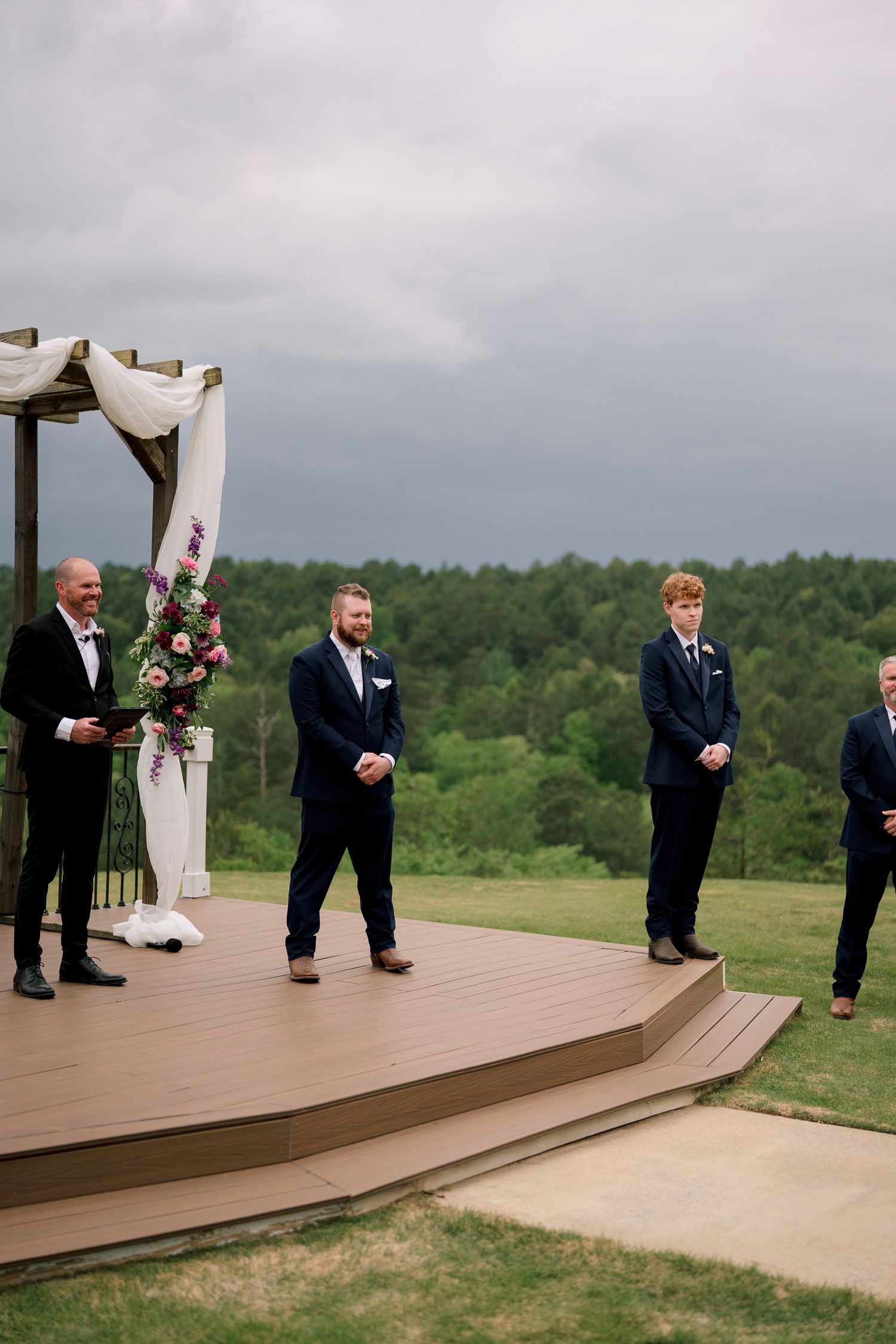 Groom and groomsmen in navy suits stand on outdoor wedding ceremony stage with floral arch and green landscape backdrop.
