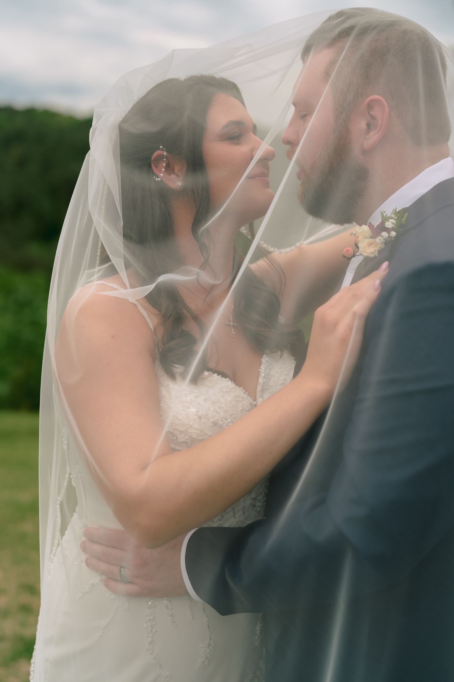 Romantic bride and groom sharing tender moment beneath flowing bridal veil with green landscape behind.