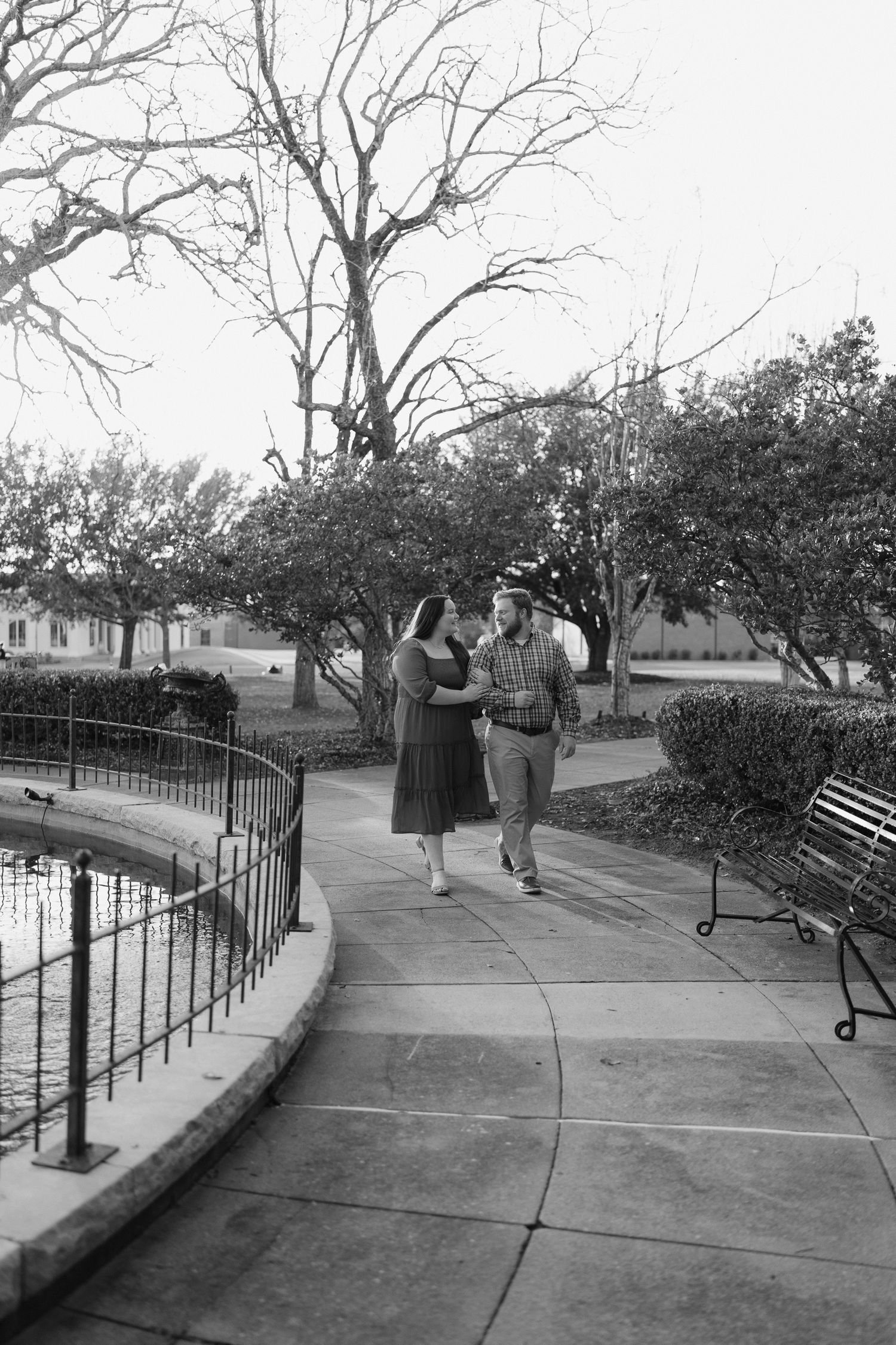 Black and white photo of a couple walking arm in arm along a park pathway beside a decorative iron fence.