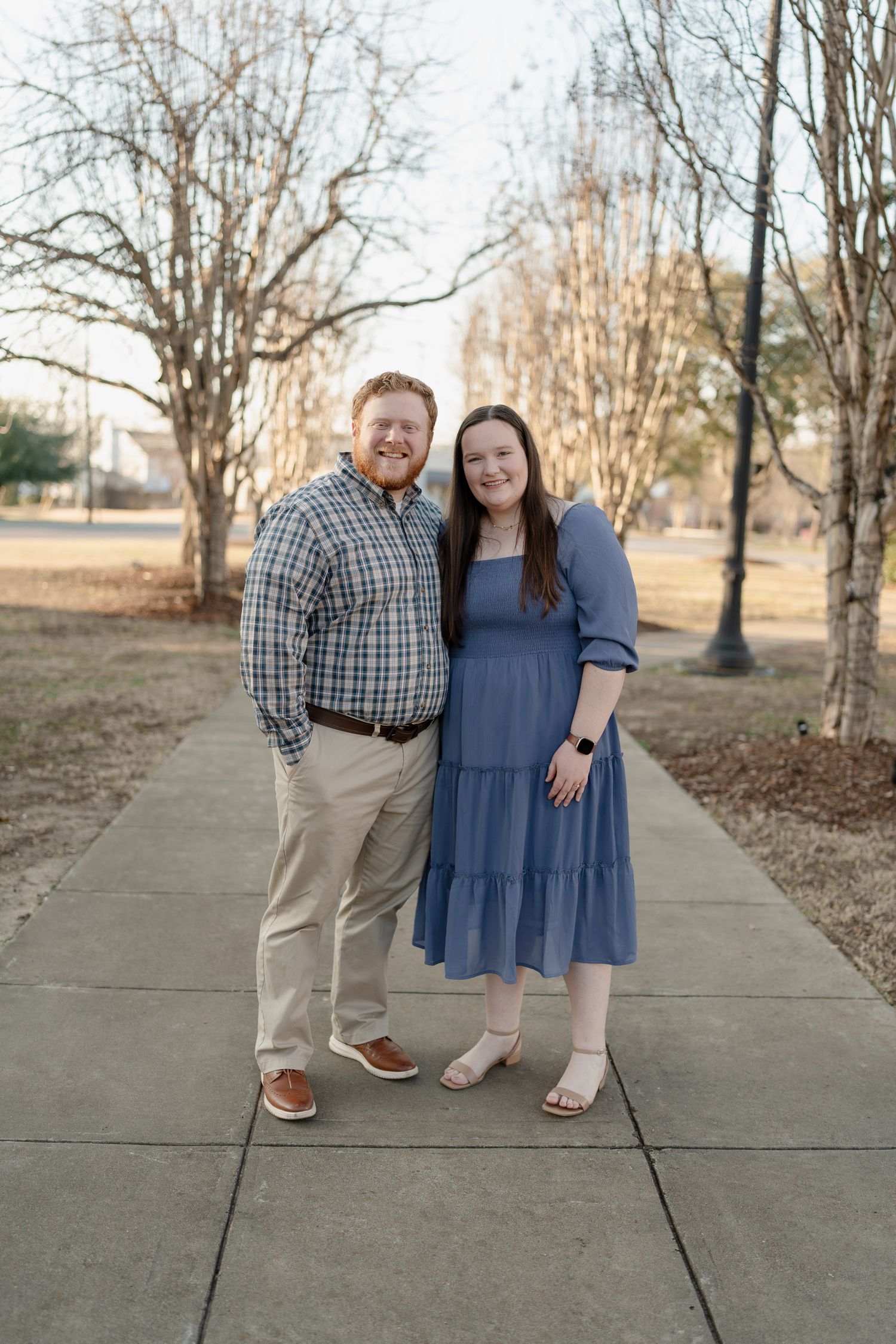 Smiling couple posing together on a sidewalk, man in plaid shirt and woman in blue dress, with trees behind.