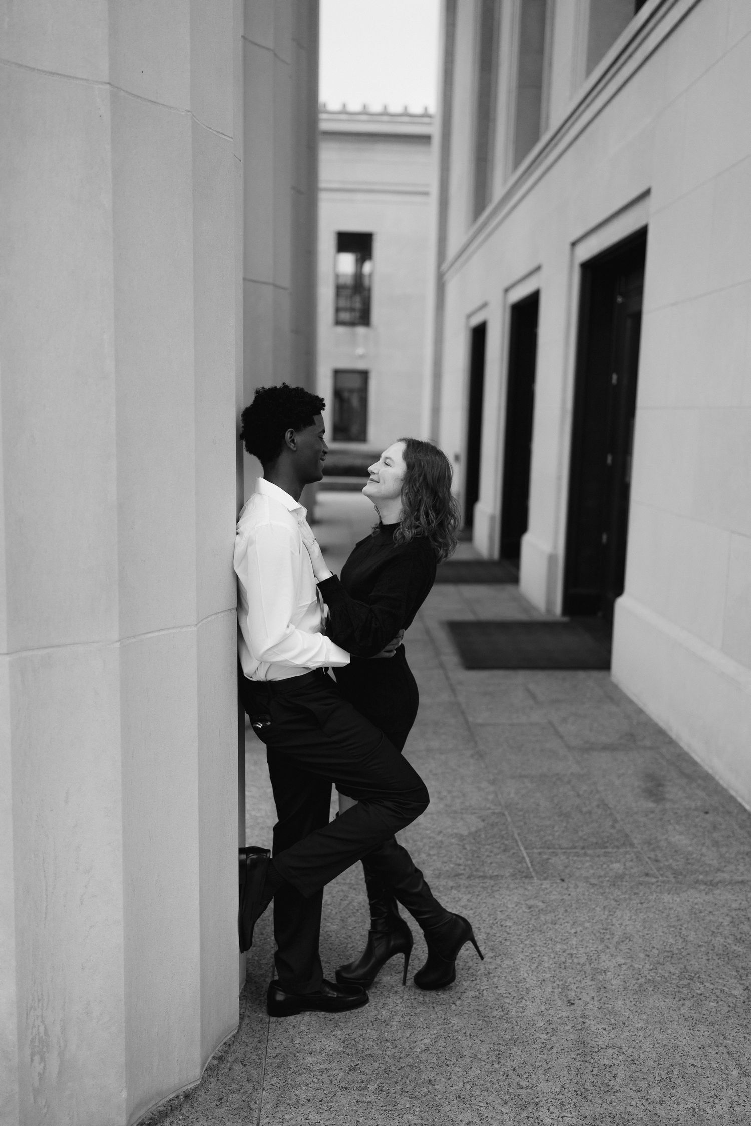 Black and white photo of interracial couple leaning against stone columns in an urban setting.