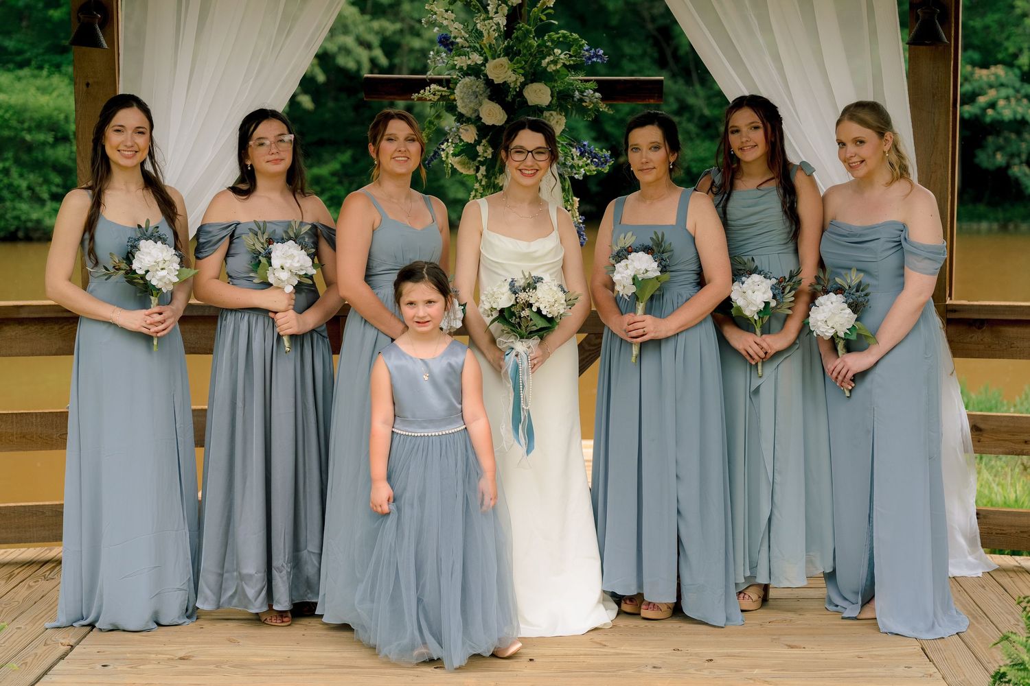 Bride poses with seven bridesmaids in dusty blue gowns and flower girl at outdoor wedding venue.
