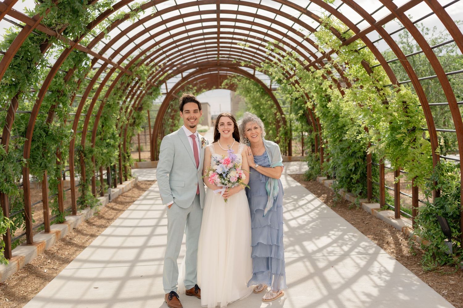 Wedding party poses under a rustic wooden archway covered in lush green vines and foliage in an outdoor garden setting.