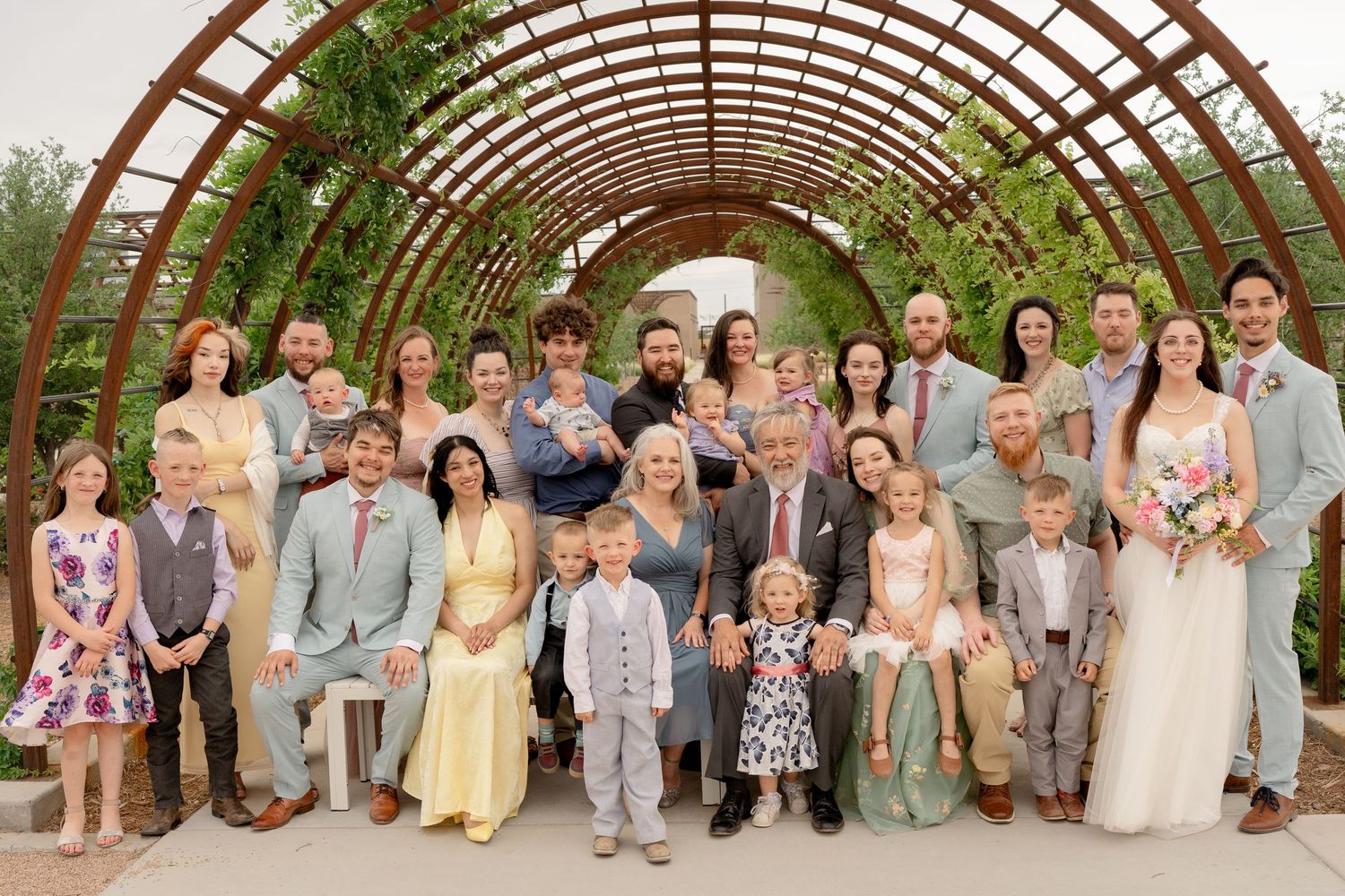 A large group photo of wedding guests and wedding party standing together outdoors under a wooden pergola.