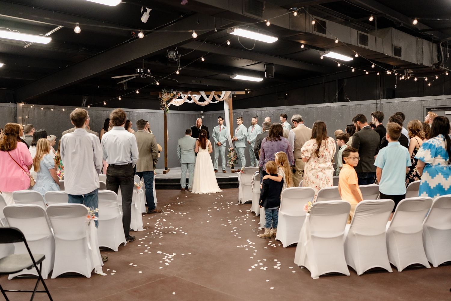 Wedding reception venue with white chair covers and scattered flower petals on dark floor.