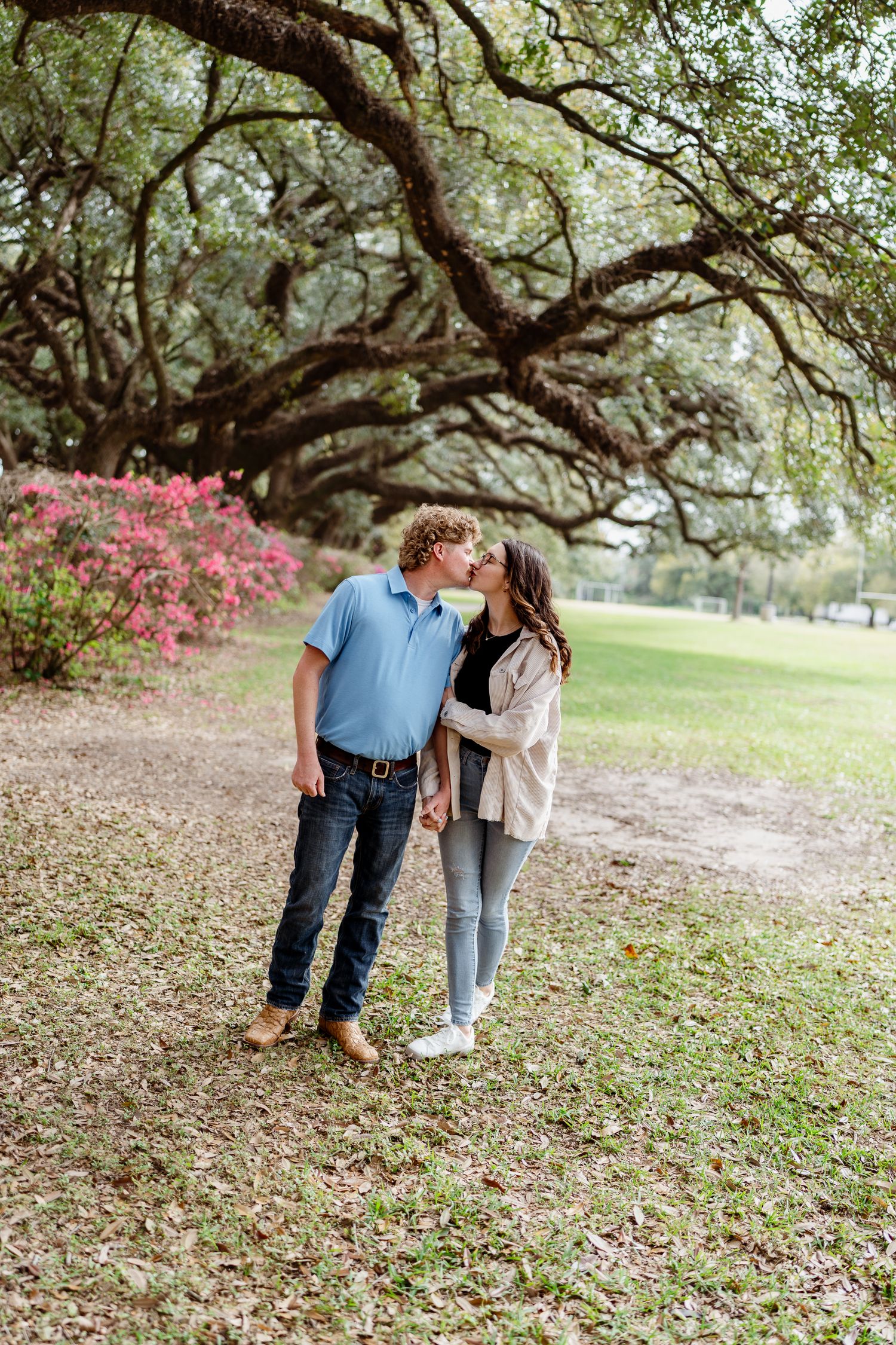 A couple walks together under oak trees on grass with pink azalea flowers blooming in the background.