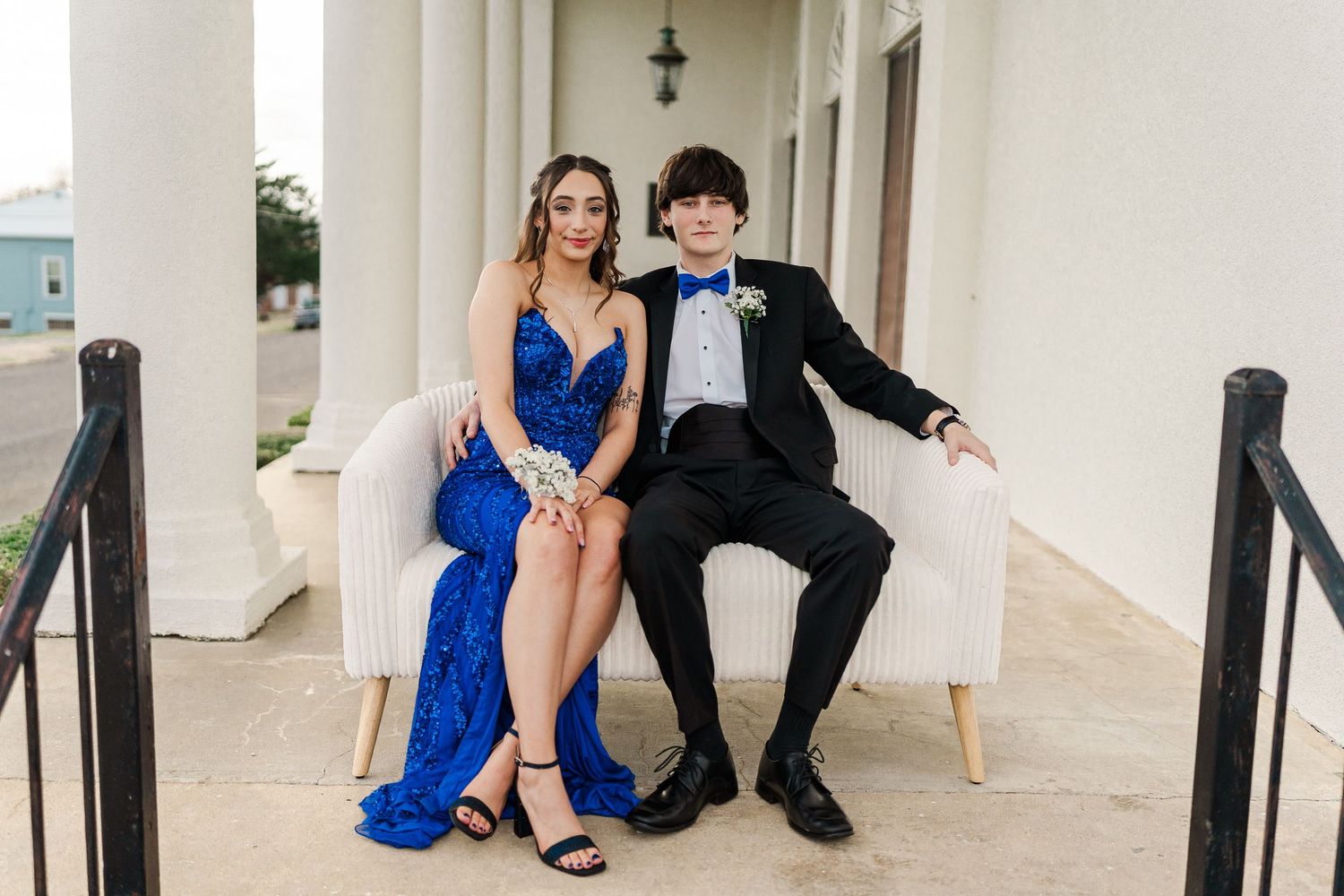 Formal couple in blue dress and tuxedo sitting together on white bench between classical columns.