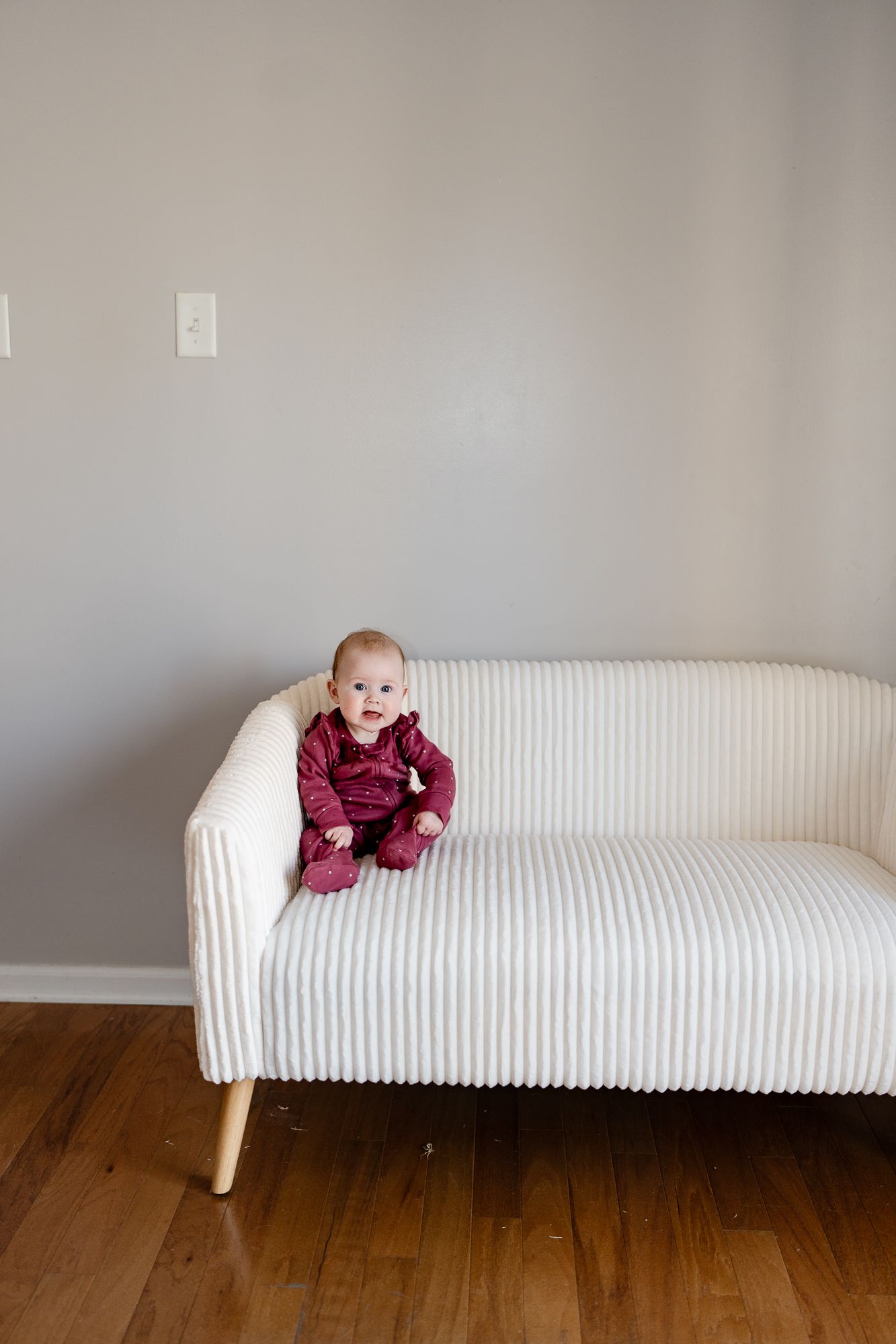Baby in burgundy outfit sits alone on white ribbed loveseat against gray wall.