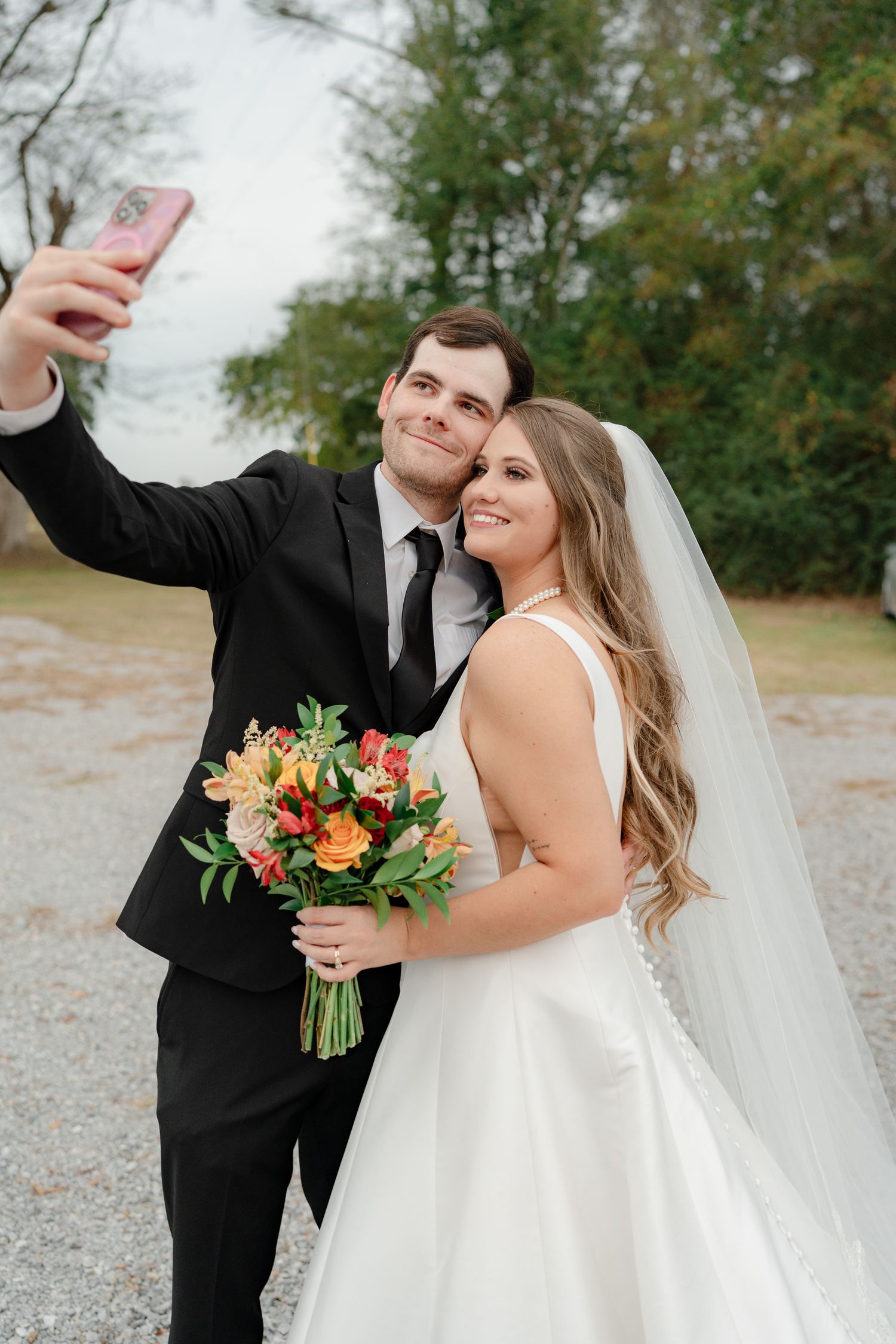 A bride and groom share joyful moments together outdoors while holding a vibrant orange and pink floral bouquet.