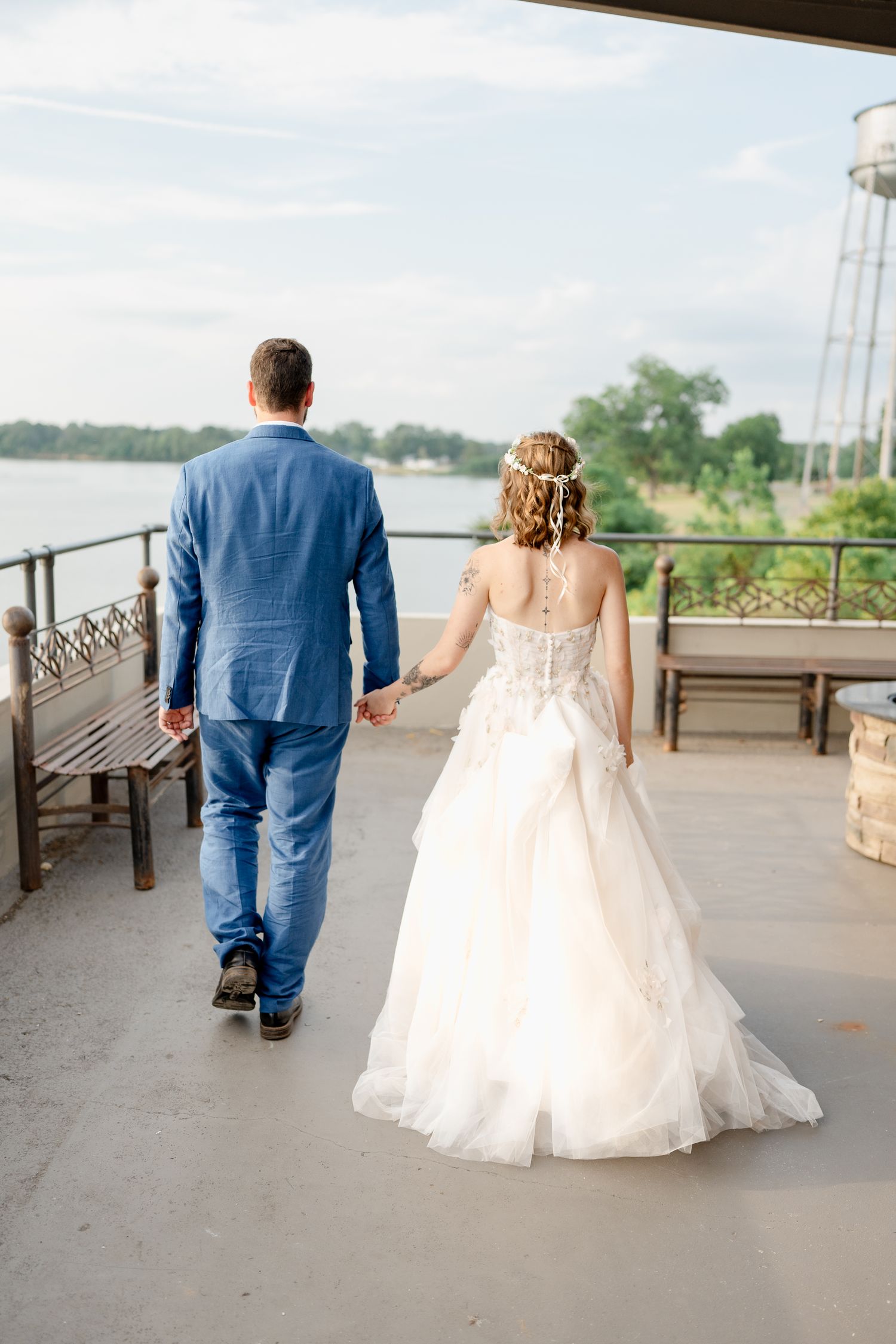 A couple walks along a waterfront promenade with the bride wearing a flowing white gown and groom in a blue suit.