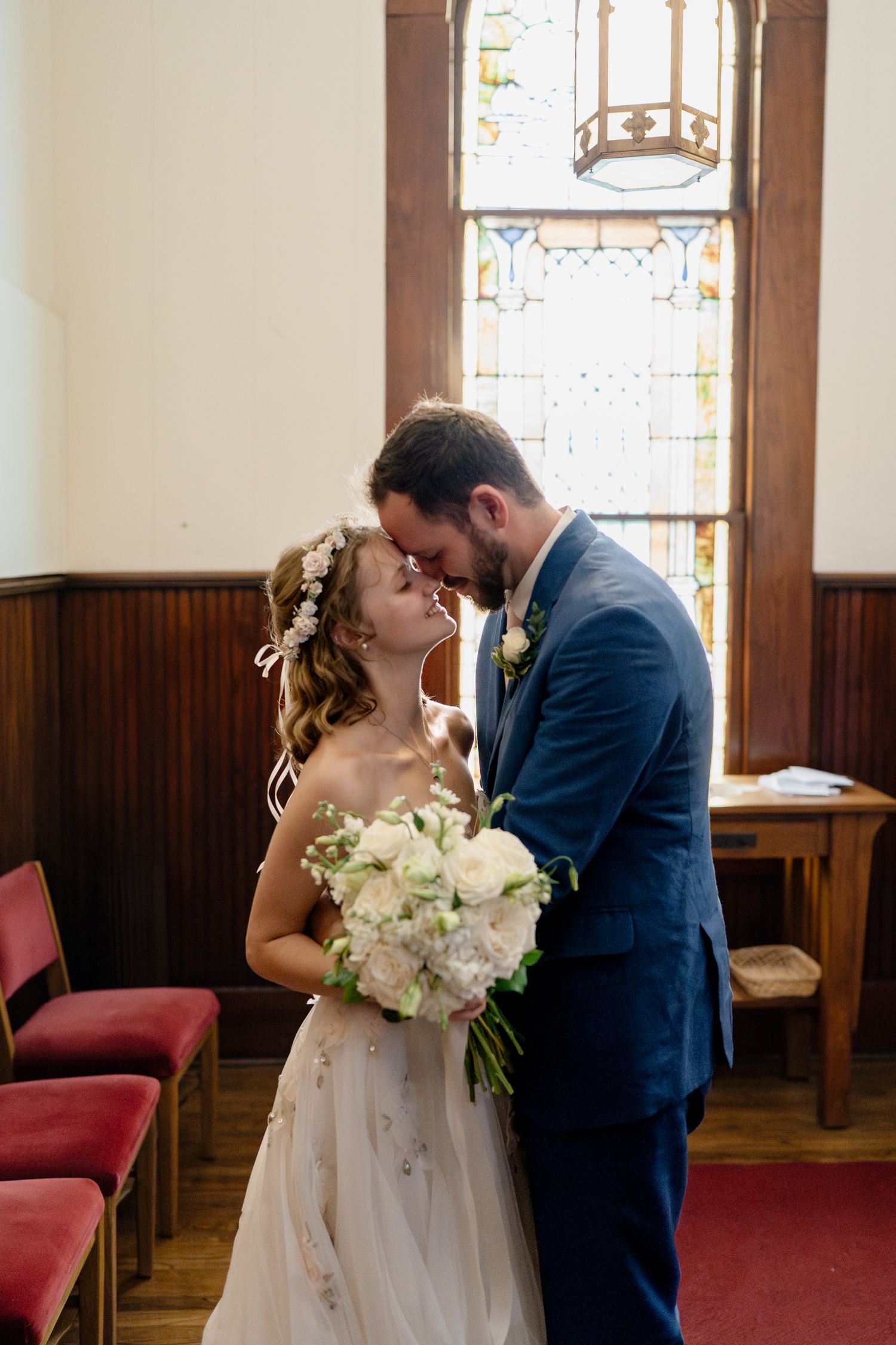 A couple shares their first kiss as newlyweds in a church with stained glass windows and wooden paneling.