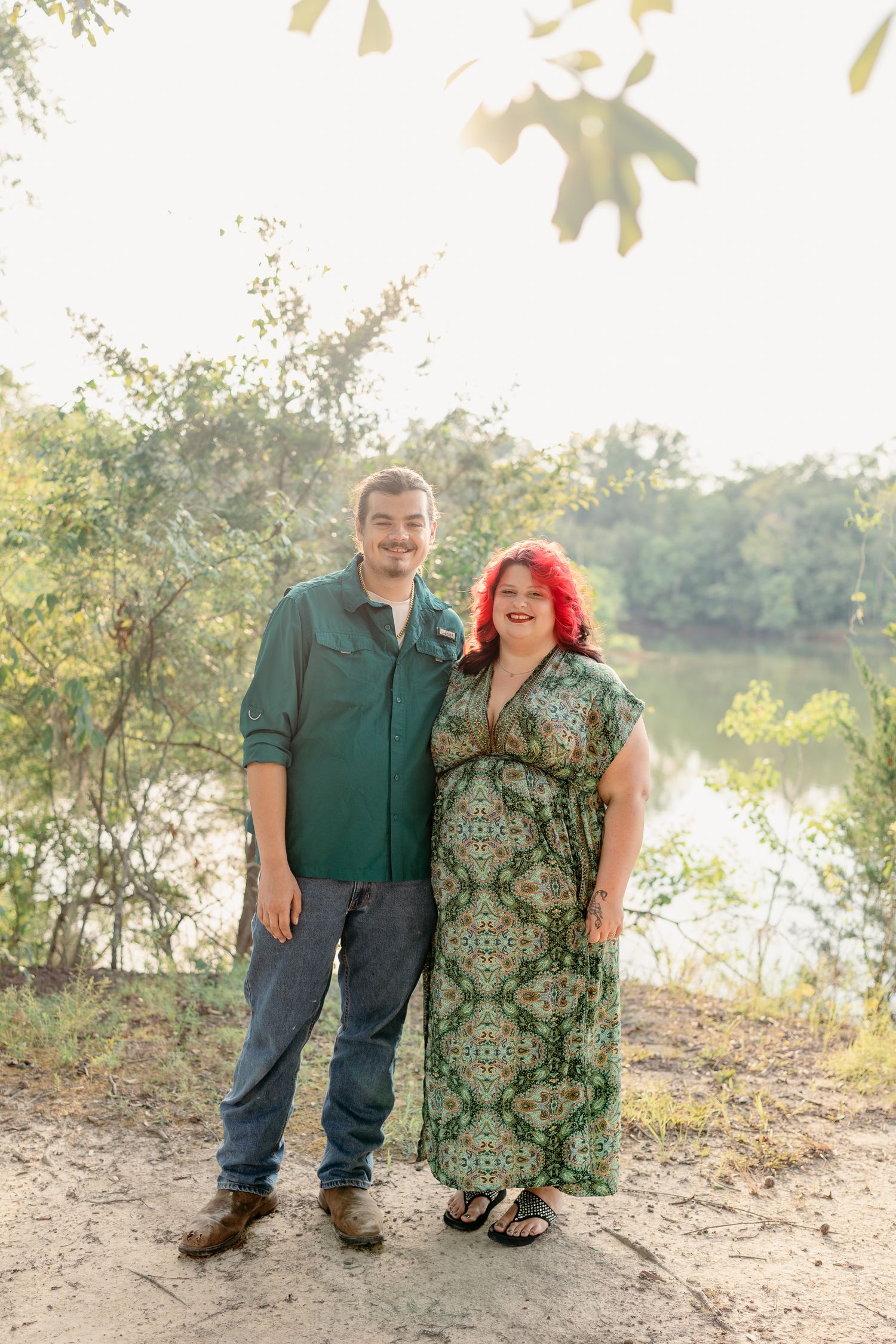 A couple stands together by a riverbank on a sunny day wearing casual clothing against a natural backdrop.