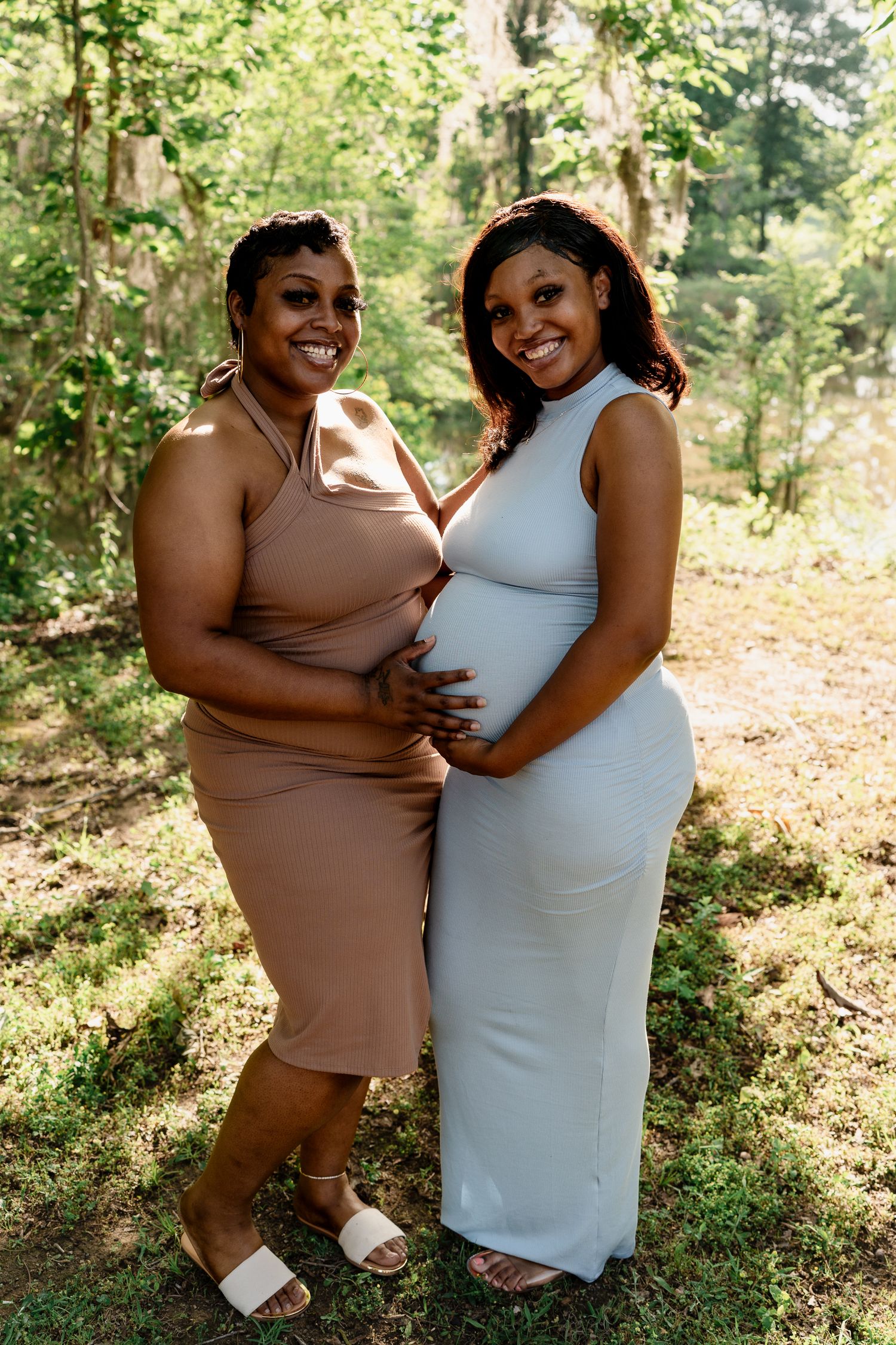 Two friends in form-fitting dresses share a joyful moment in a sunlit forest.