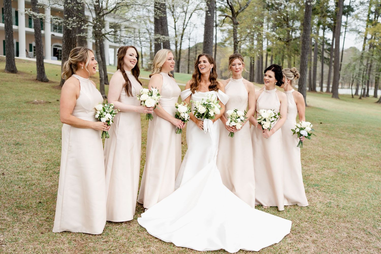 Bridal party poses outdoors in coordinating cream dresses holding white floral bouquets.