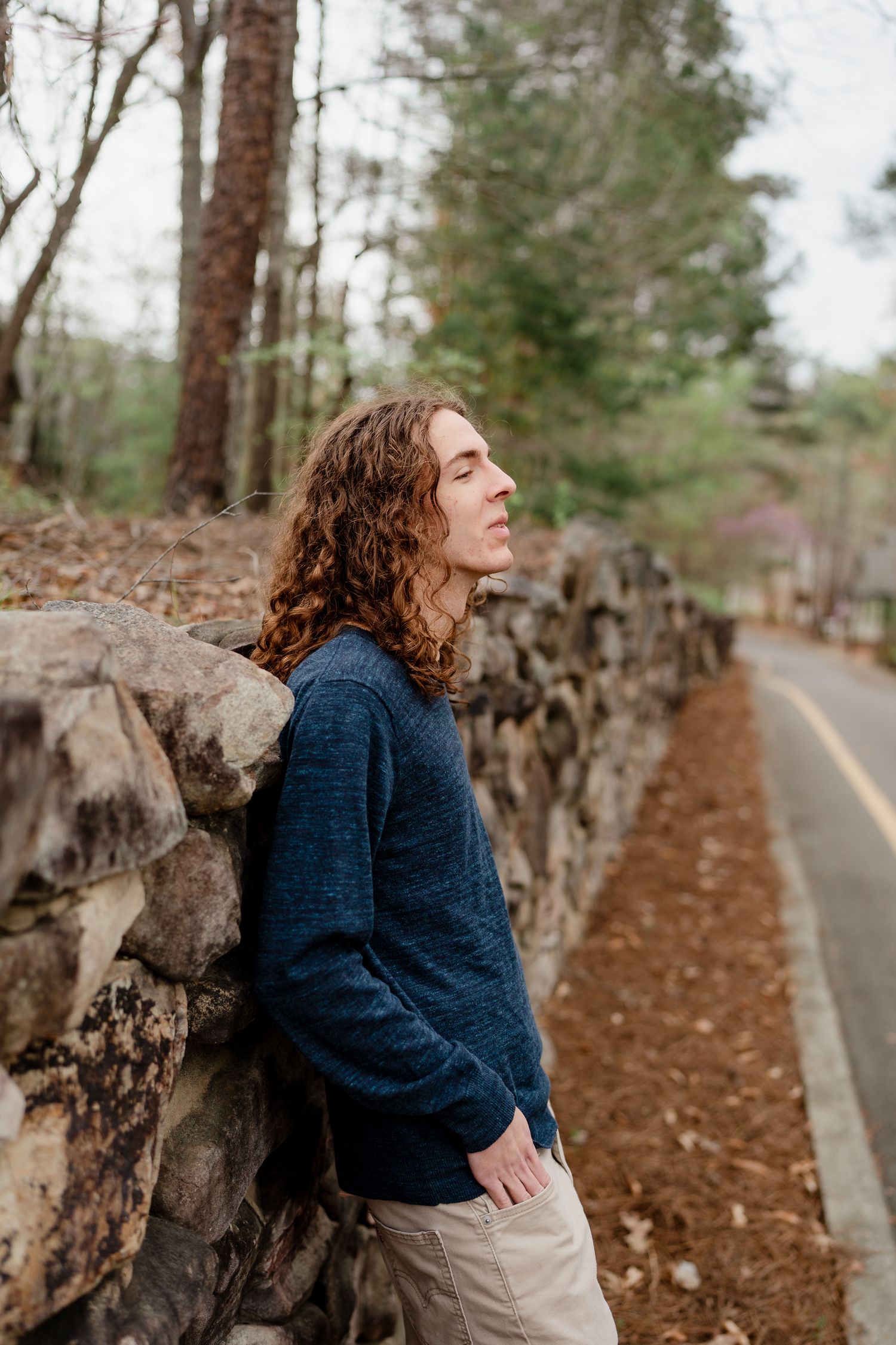Person in navy sweater leaning against stone wall on tree-lined path during autumn.