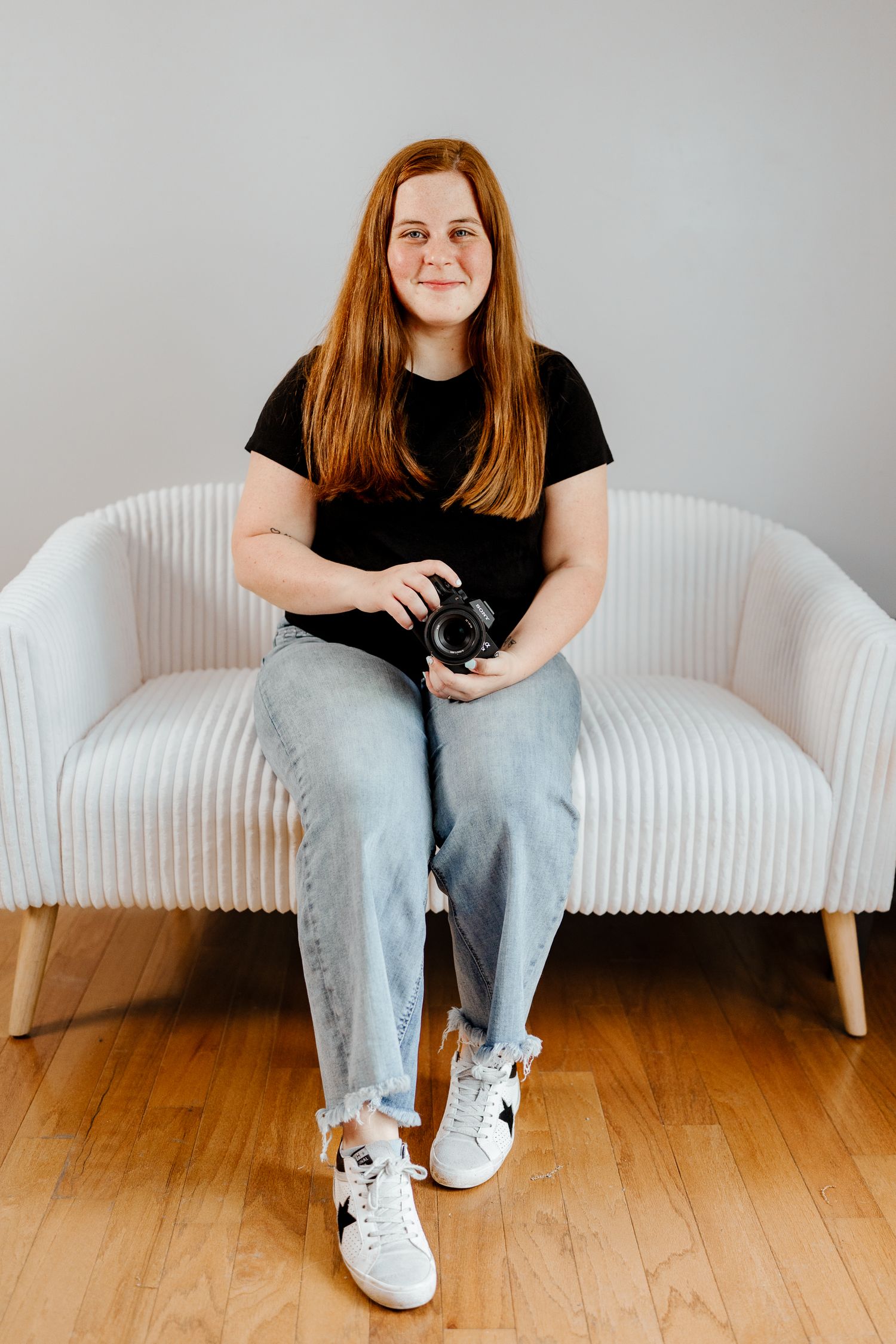 Casual indoor photoshoot featuring person in black top and jeans on white textured couch.