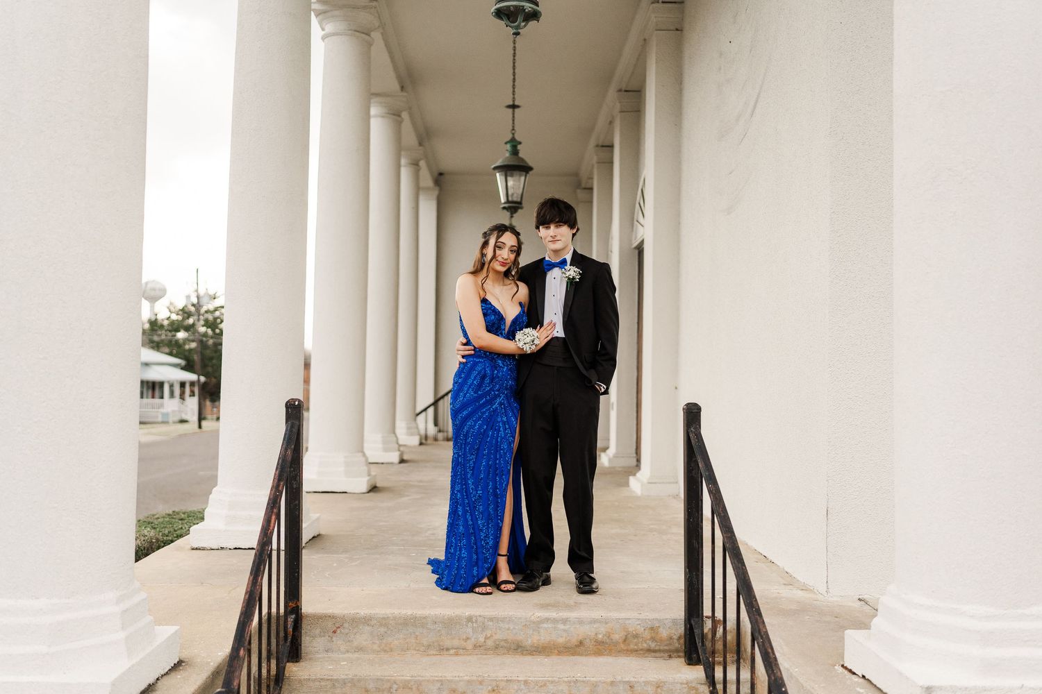 A couple in formal attire poses in an elegant white columned walkway for a special occasion photo.