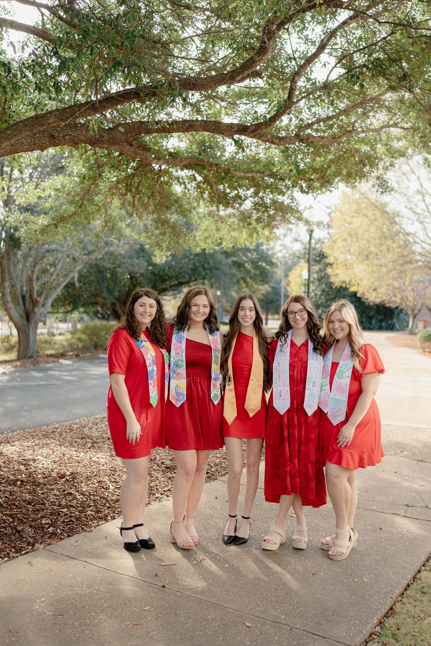 A group wearing matching red dresses and white scarves poses together under trees on a path outdoors.