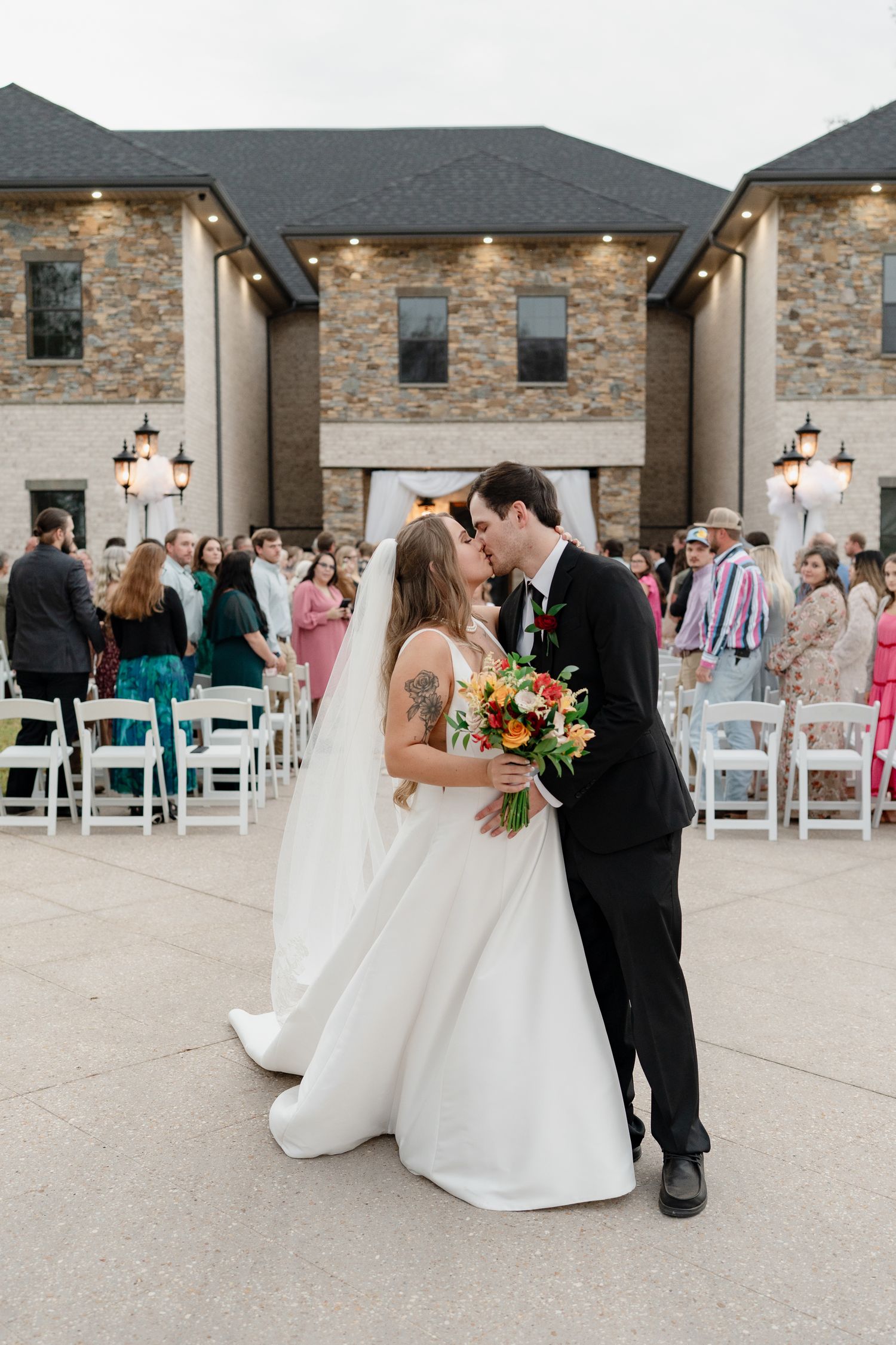 Newlyweds share their first kiss outside a stone venue as wedding guests look on.