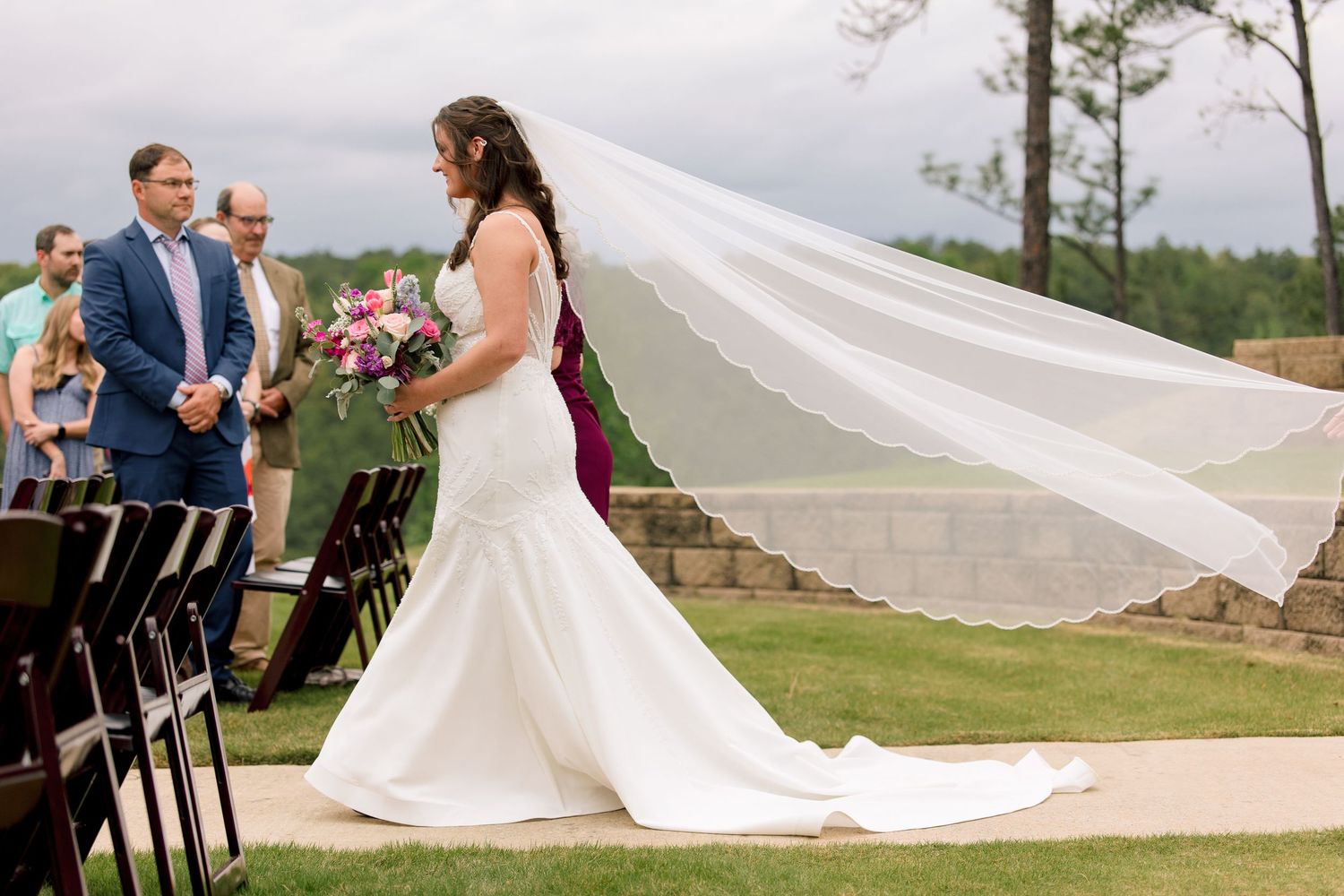 Bride in white mermaid gown with veil blowing in wind walks down outdoor wedding aisle holding pink bouquet.