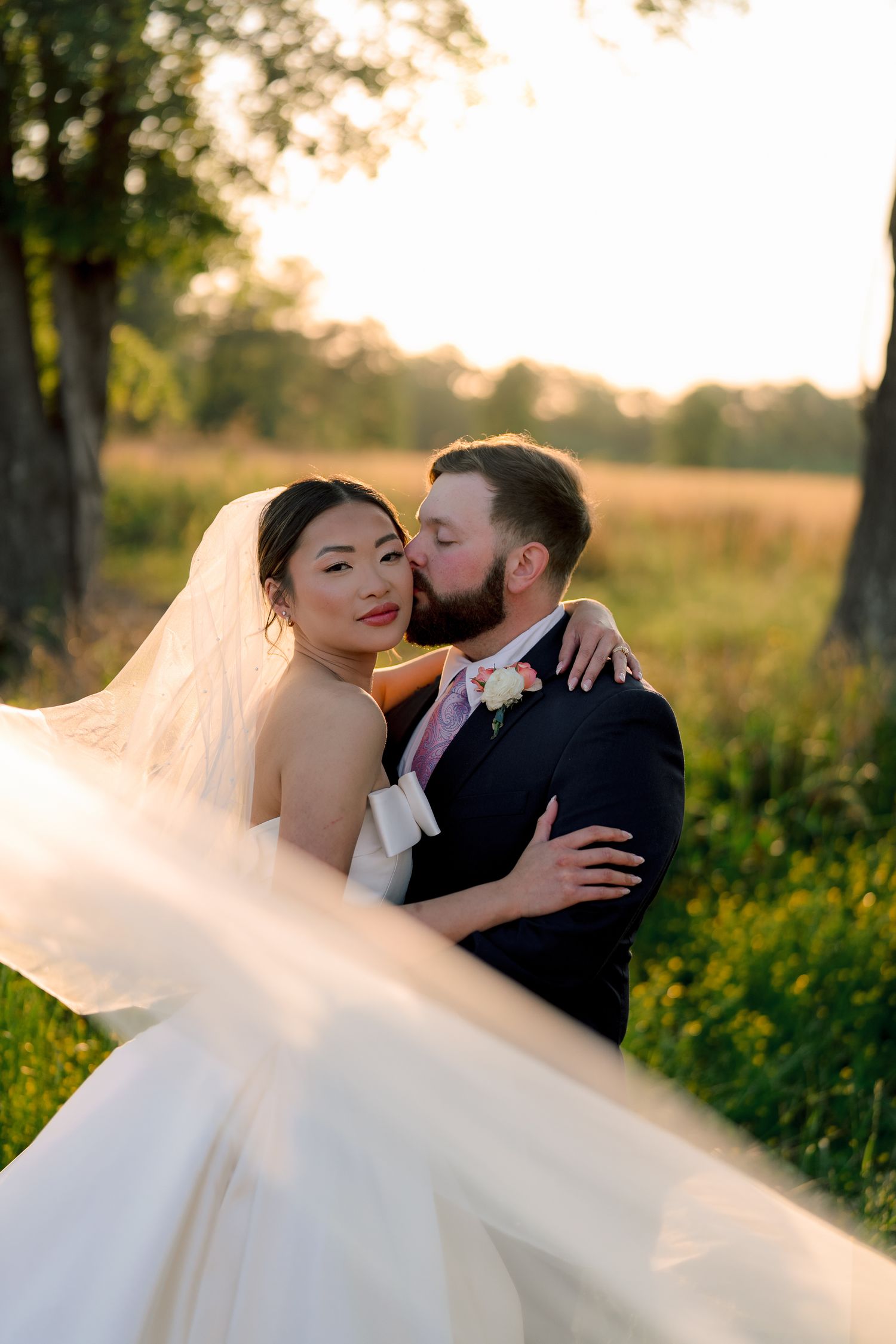 A bride and groom share a romantic sunset kiss outdoors, her white veil flowing in the breeze amid lush green fields.