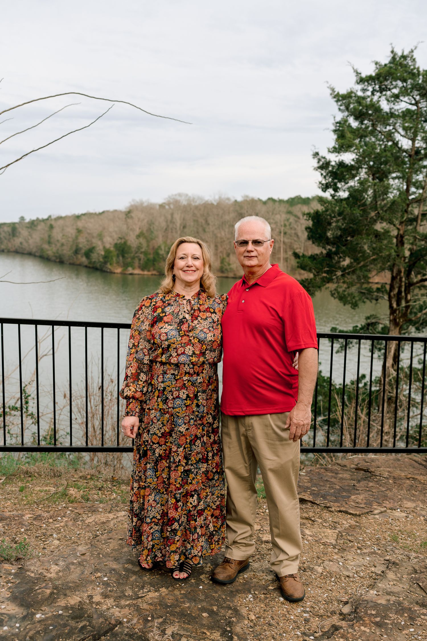 Couple posing together by a railing with a scenic river and wooded landscape behind them.