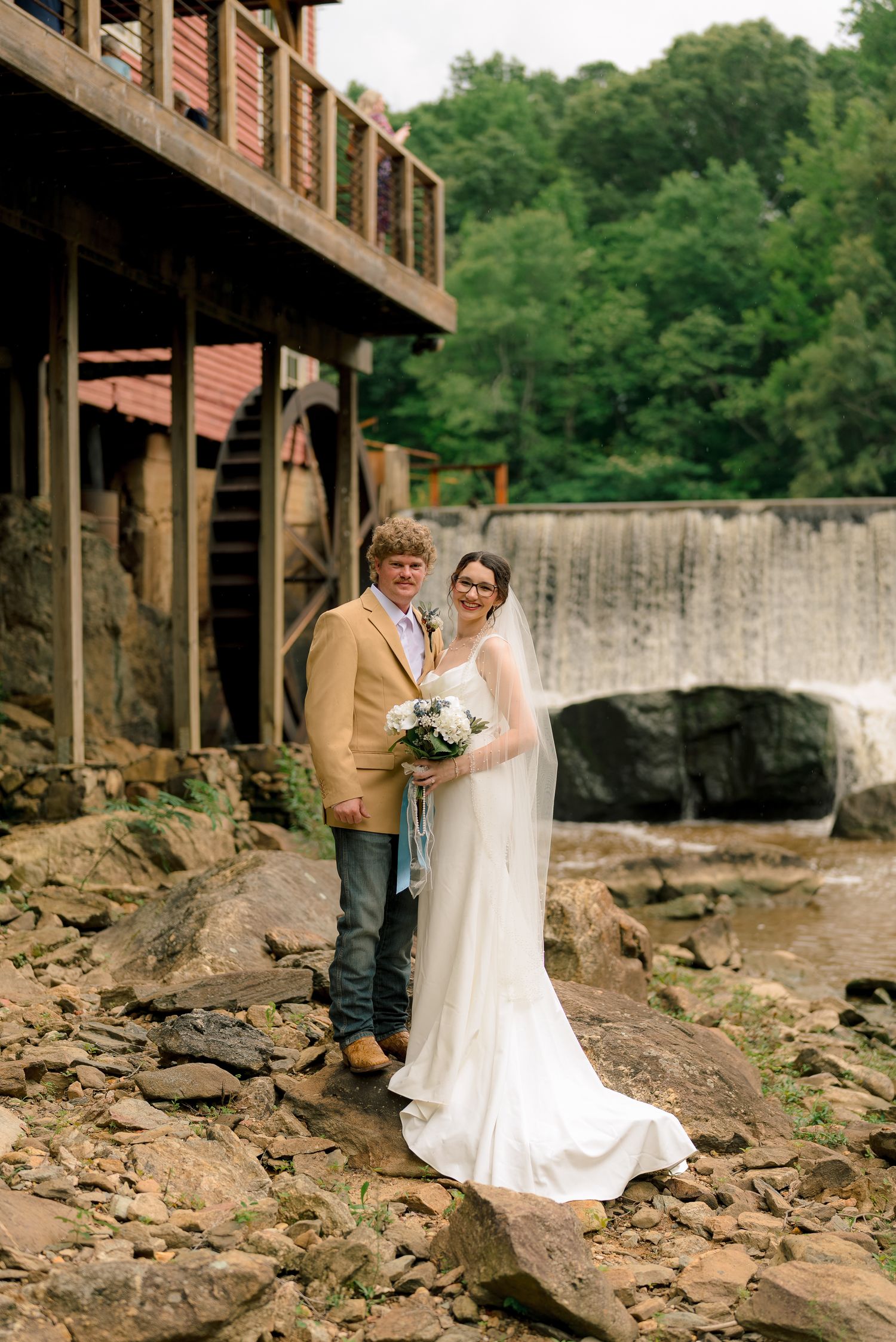 Smiling bride and groom pose on rocky riverbank beside a waterfall and rustic mill building.