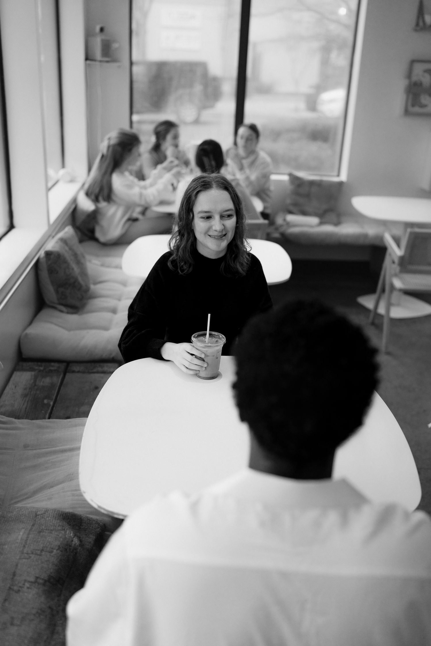 A smiling woman in black holds an iced drink across a white table from a man in a cozy cafe setting.