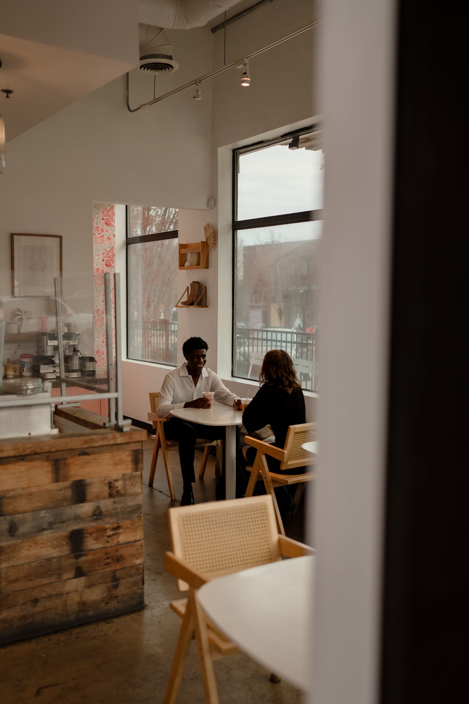 Two people sit across from each other at a small table in a cozy cafe with large windows.