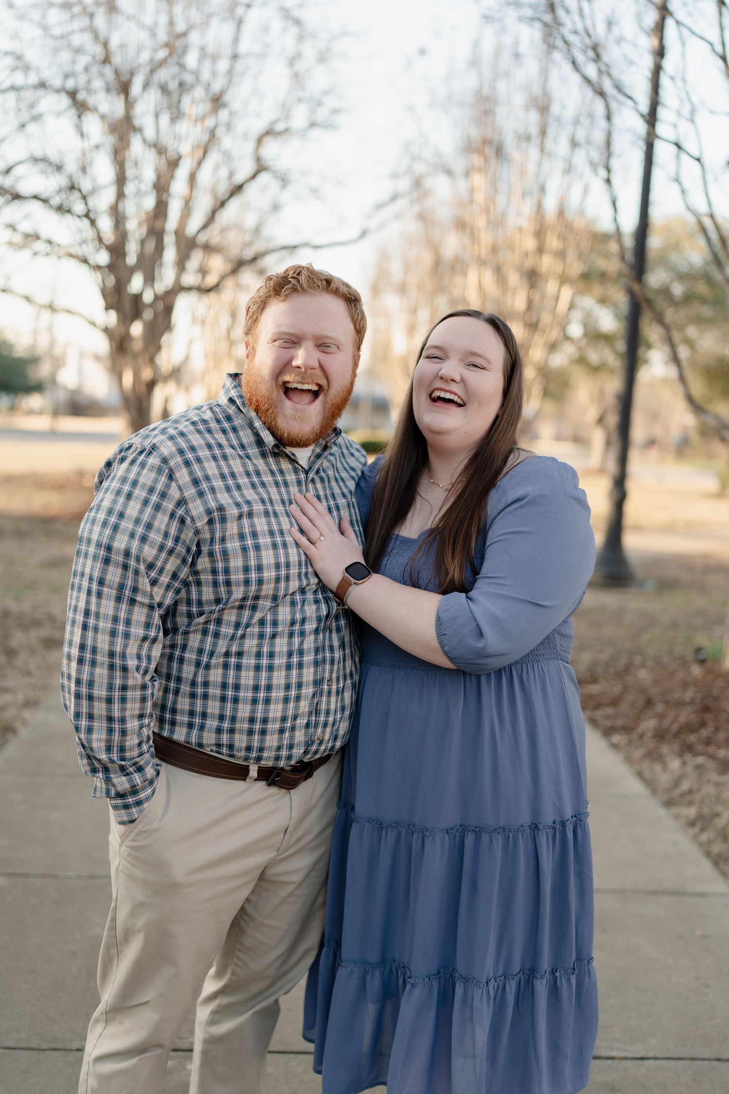Laughing couple poses outdoors on a sidewalk, man in plaid shirt and woman in blue dress, sharing a joyful moment.
