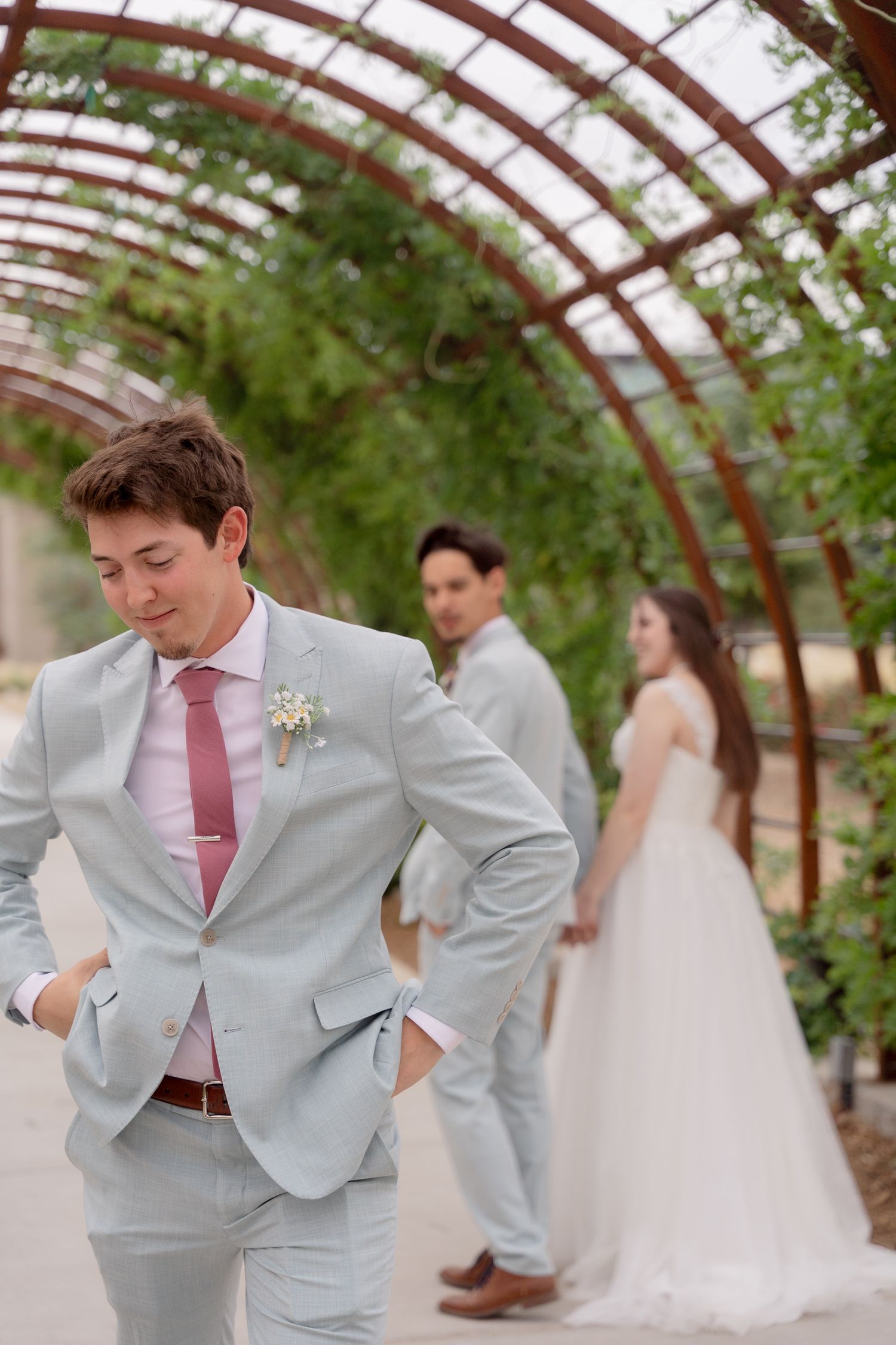 Wedding party members in light gray suits and pink ties pose in a rustic greenhouse venue.