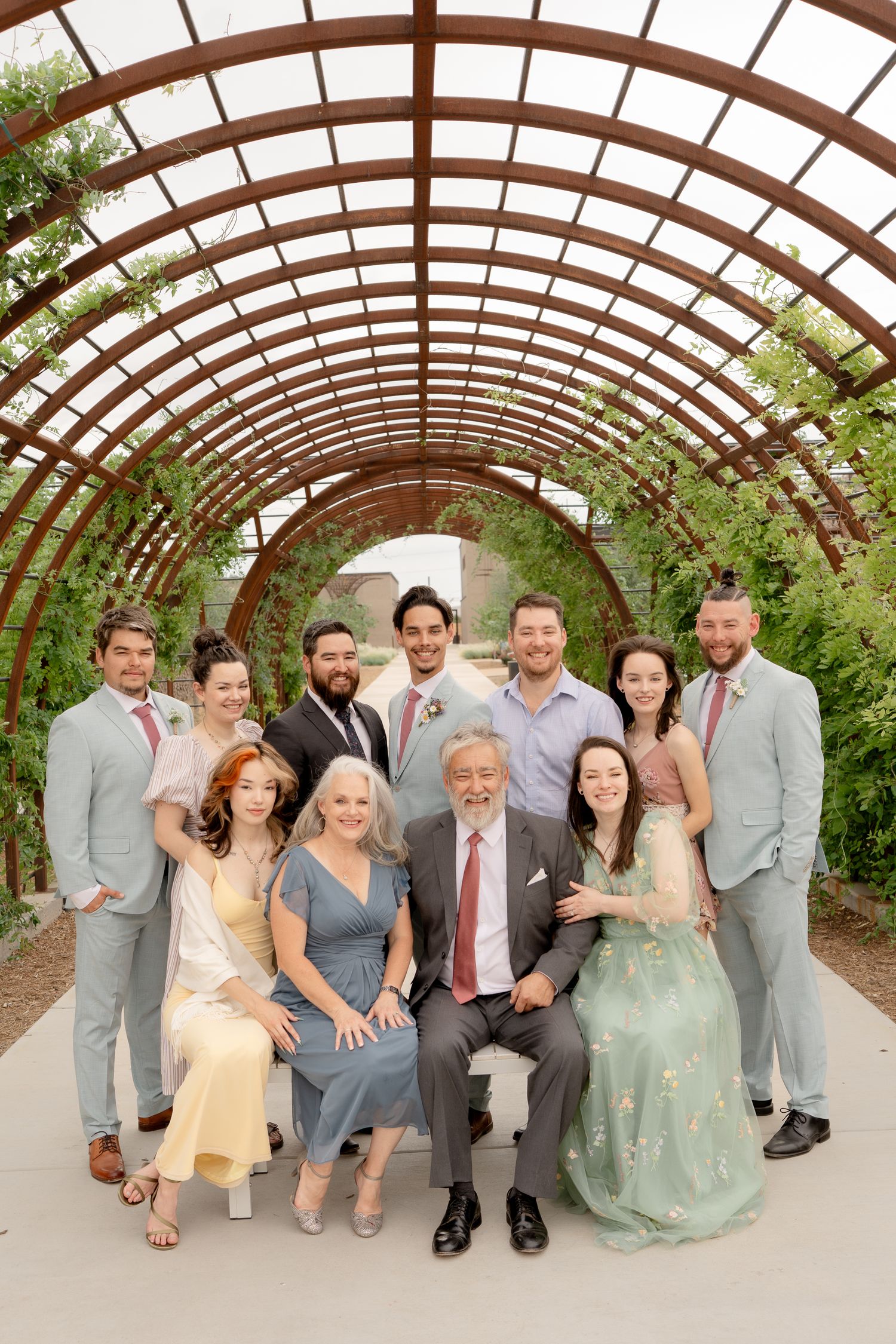 Wedding party poses together under an arched garden pergola with wooden beams and climbing vines.