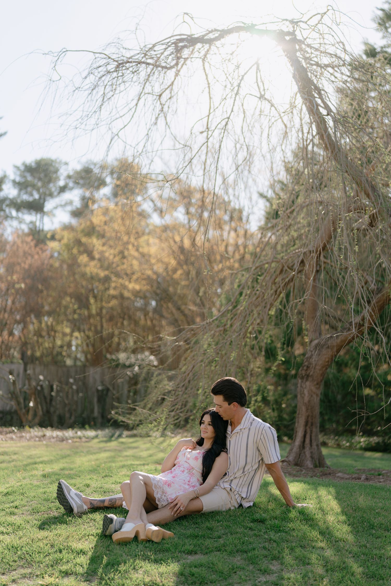 Couple sitting together on green grass under a bare weeping tree in a sunlit park during fall.