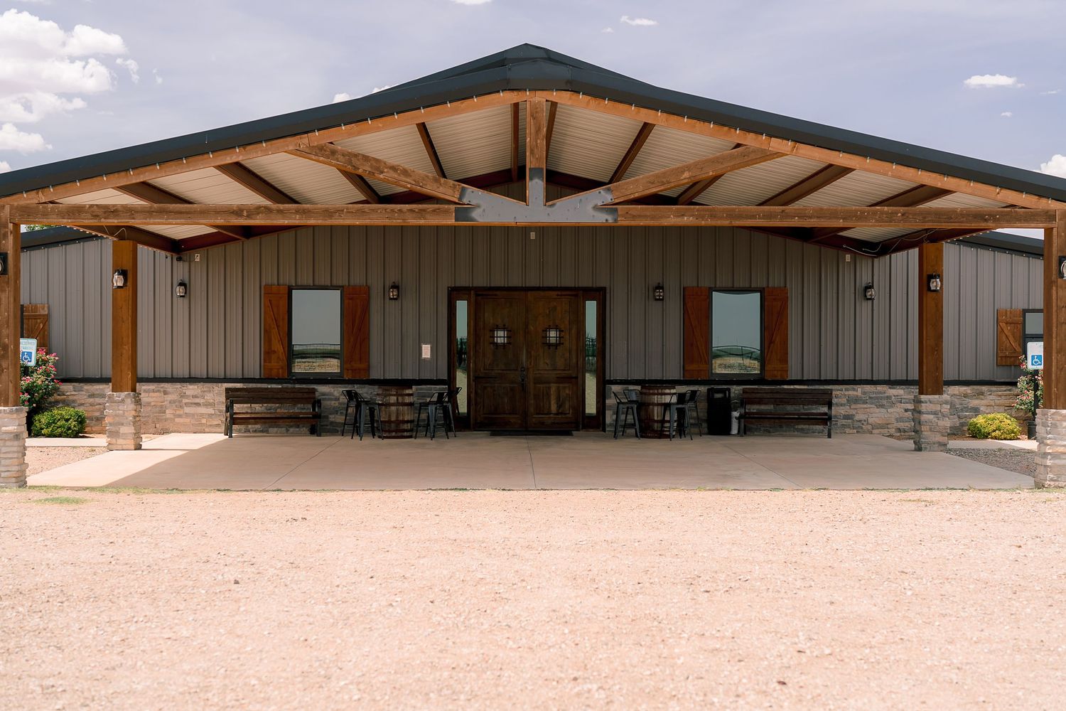 Rustic event venue with wood beam entrance, double doors, outdoor seating, and metal siding under a partly cloudy sky.