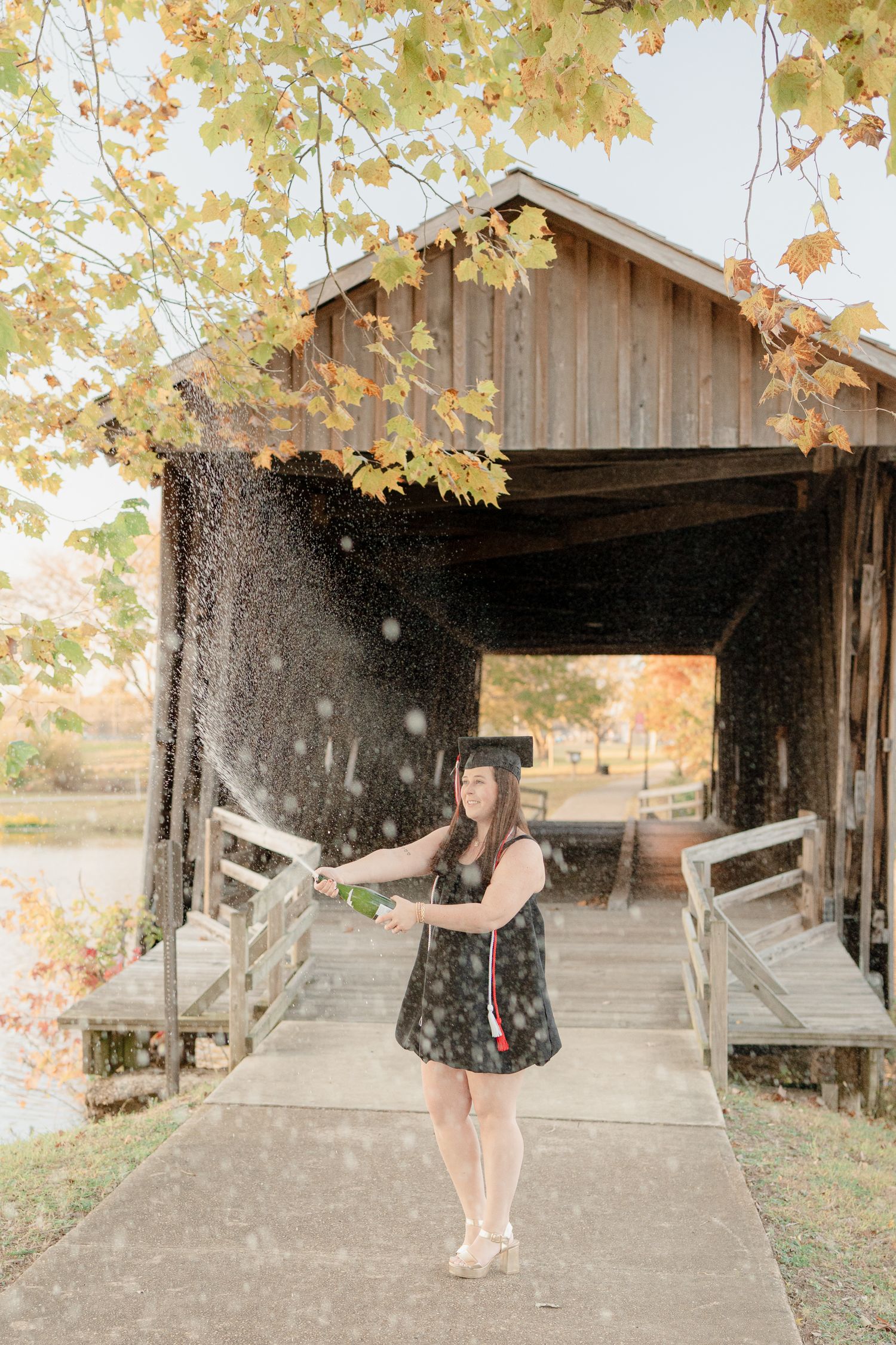 Graduate in cap and gown twirls joyfully under autumn leaves at a covered wooden bridge.