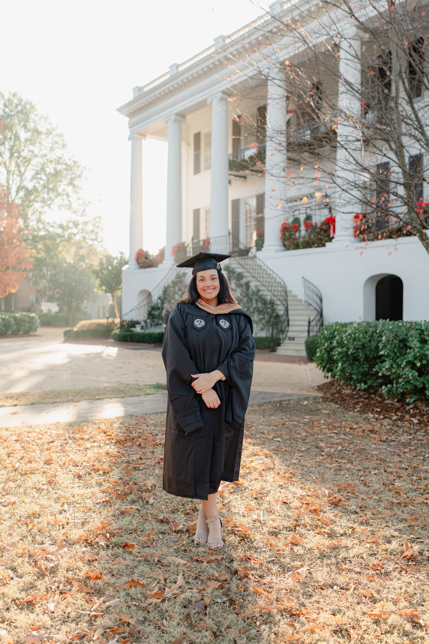Graduate in cap and gown poses in front of stately white columned building with autumn leaves on ground.