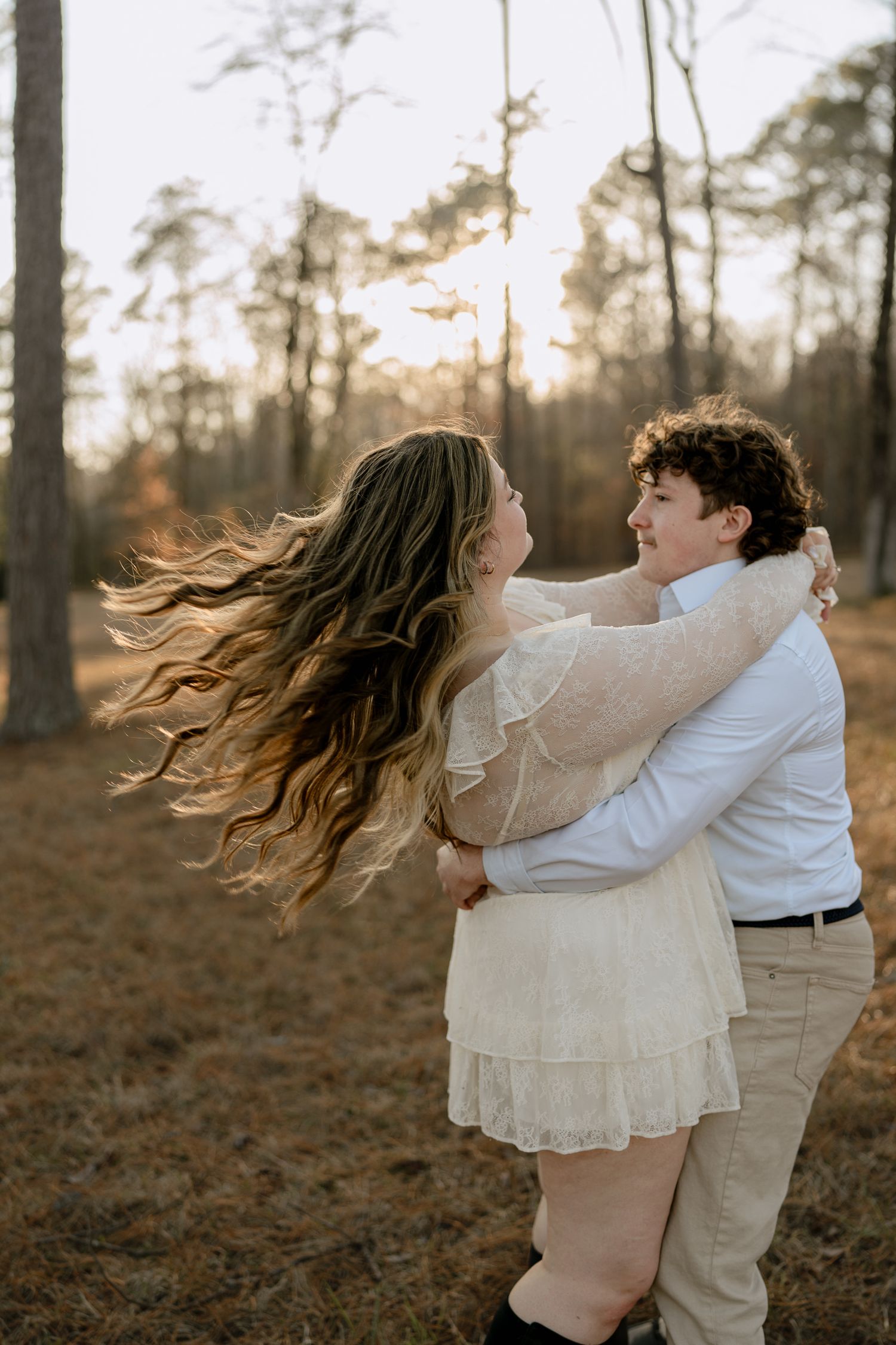 Couple embracing and laughing in a golden-hour pine forest, woman's long hair flowing in the breeze.