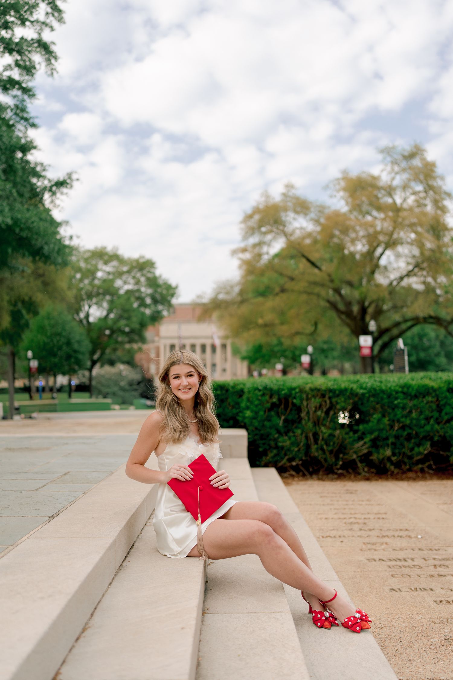A person in a white dress sits on concrete steps holding a red heart-shaped object and wearing red polka dot shoes.