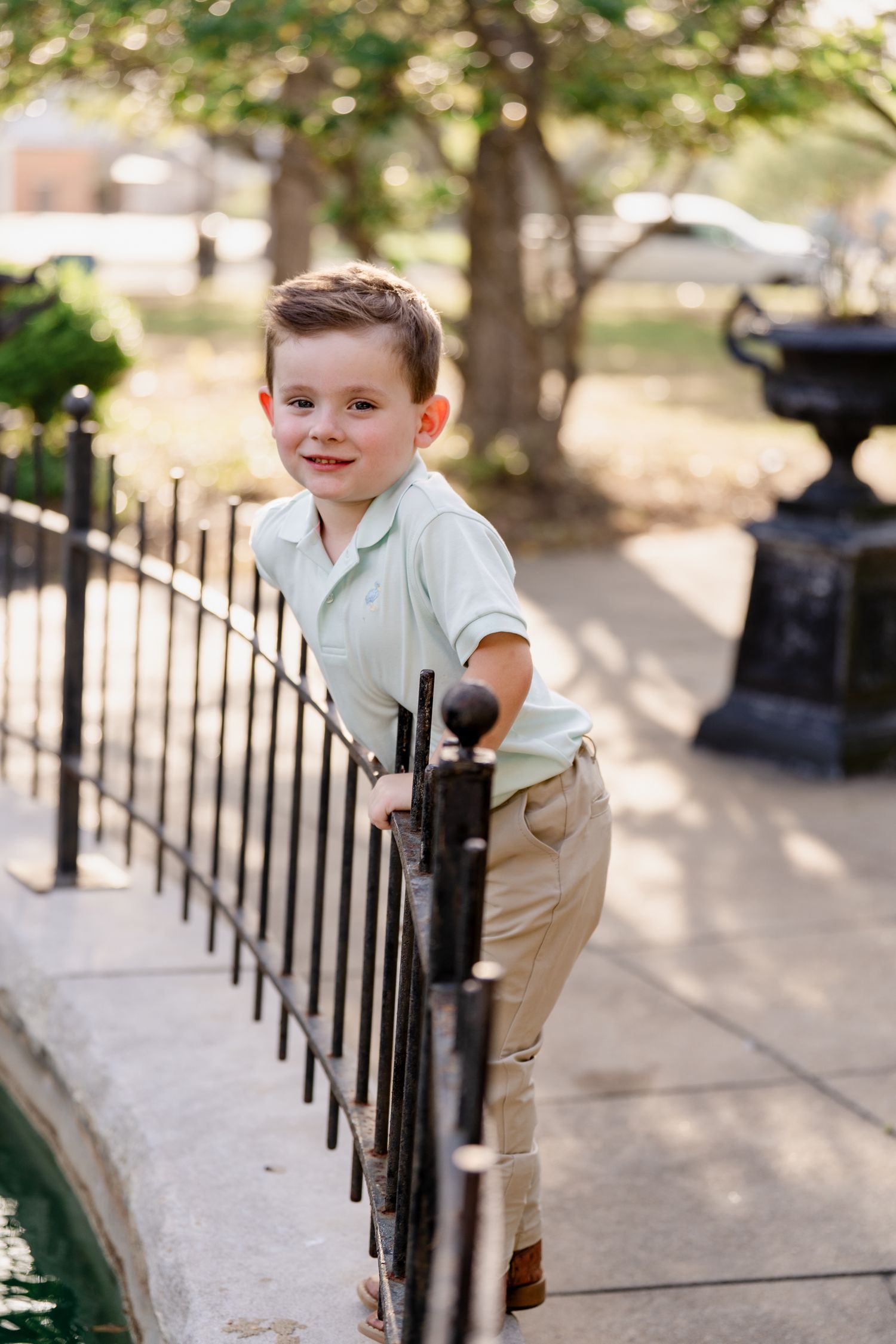 A young child in light colored clothing leans playfully against an iron railing near water.